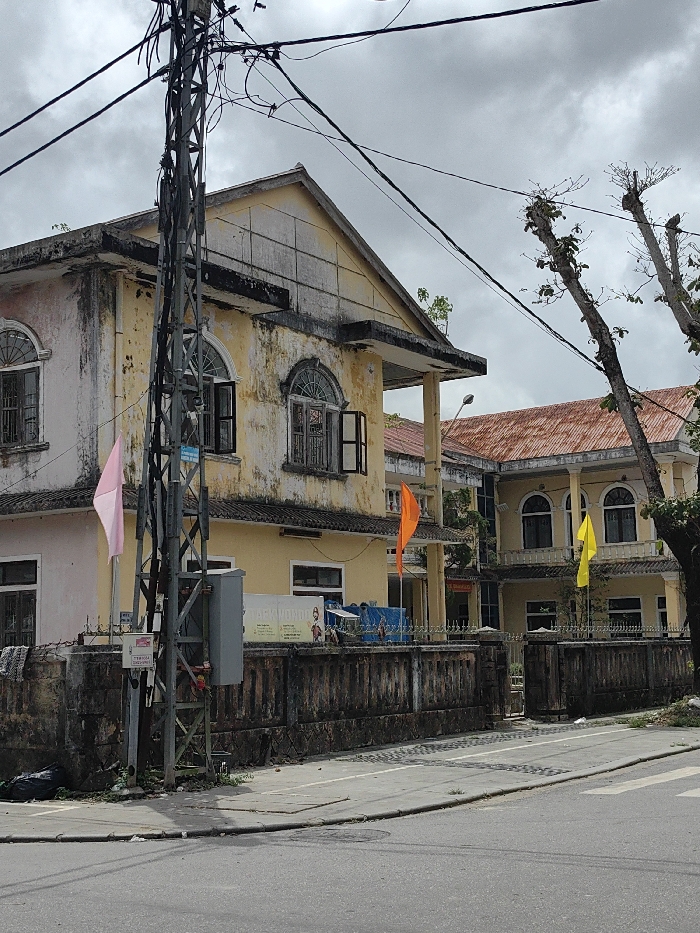 The image presents an urban street scene in Phú Xuân Ward, Vietnam, under an overcast sky. In the foreground, an asphalt road leads to a concrete sidewalk bordered by a curb. A black refuse bag is positioned on the sidewalk to the far left. A prominent, gray metal lattice utility pole, supporting numerous black electrical and communication cables that crisscross the upper portion of the frame, stands in the left-center. A gray electrical enclosure is attached to this pole.
Behind the sidewalk and utility pole, a dark, weathered stone or concrete fence with decorative ironwork encloses two multi-story buildings. The structure to the left, which is closer, features a faded light yellow and pale pink facade, exhibiting widespread staining and organic growth. It has a gabled roof section and an overhang supported by columns. Two upper-story windows with arched tops and dark-framed sashes are open. A light pink flag is affixed to a pole near the left side of this building, and an orange flag is flown further along the fence. A partially visible sign displaying "TAEKWONDO" and a martial artist graphic is mounted on the fence. Blue tarpaulins are visible behind the fence line.
Further into the scene, a second building with similar architectural characteristics, including arched windows and a yellow facade, is visible. This building is topped with a reddish-brown corrugated metal roof. A yellow flag is flown near this structure. A street light fixture is positioned between the two buildings above the fence line. To the right, several deciduous trees with sparse new leaf growth stand against the sky. No individuals are present in the depicted scene.