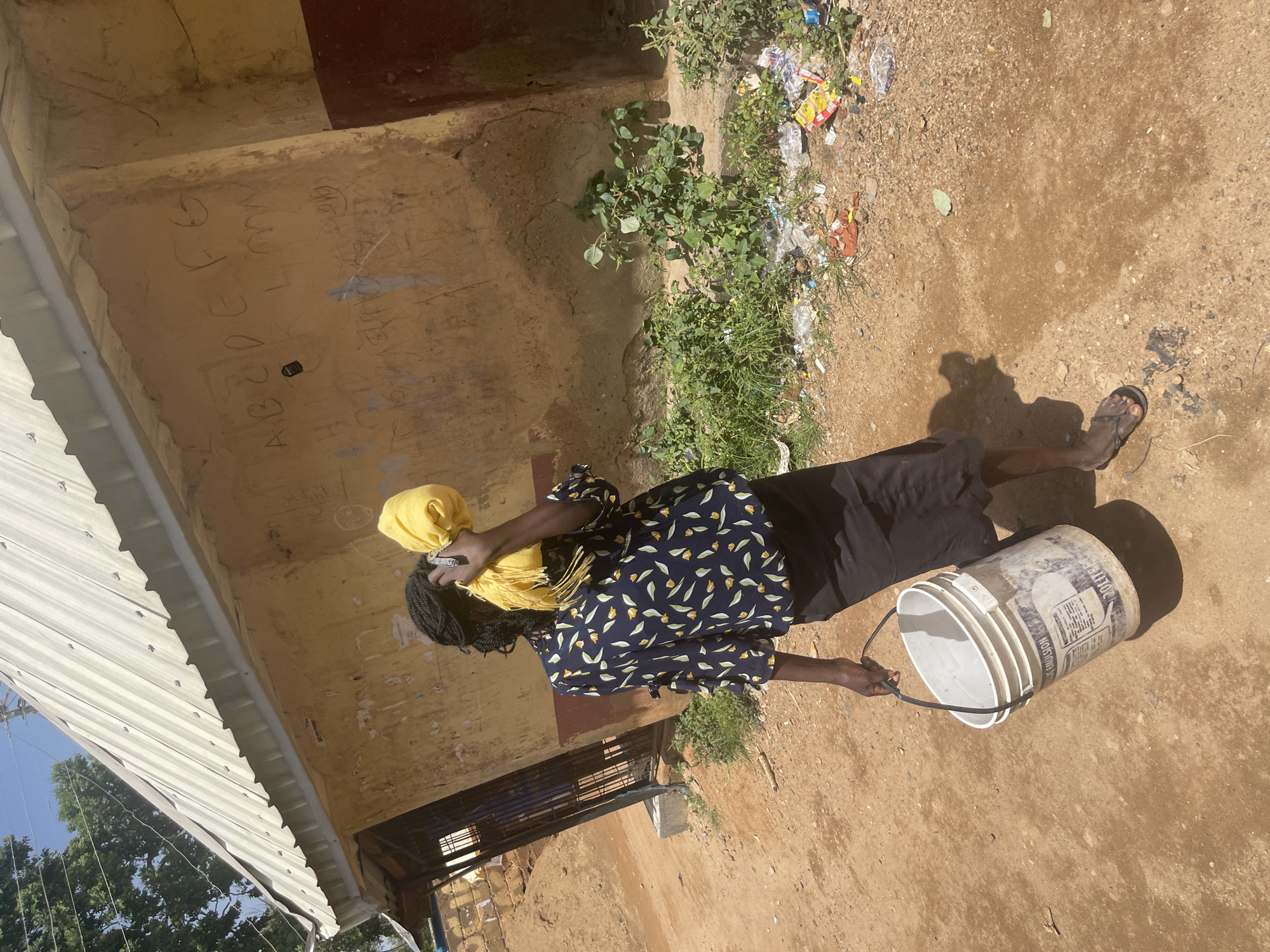 A person is standing on a dirt path outside a rough, yellow-walled building in Jos, Nigeria. The person is facing away from the camera, holding a yellow cloth over their face and carrying a white bucket with a black handle in their right hand. They are wearing a dark blue shirt with a floral pattern and a dark skirt. The building's wall has graffiti, including what appears to be the letters "A B C D E F G" and other scribbled markings. There are some green plants and scattered debris, including plastic bags and wrappers, on the ground near the building. A corrugated metal roof is visible on the building. The scene is brightly lit by daylight, casting shadows on the ground.