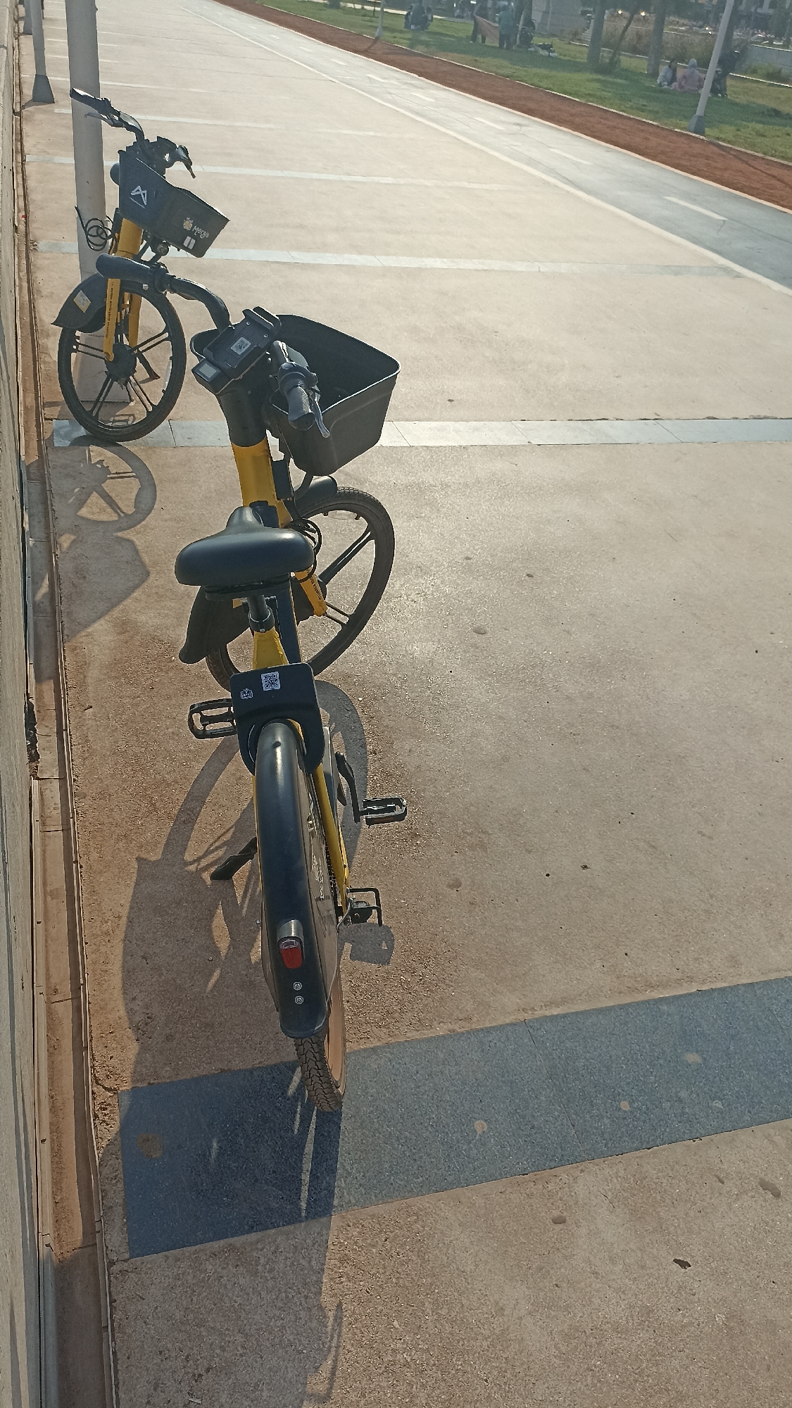 Two yellow and black rental bicycles are parked on a concrete sidewalk. The bicycle in the foreground has its back wheel to the camera, and its front is angled to the left. It has a black basket on the handlebars, a black seat, and black fenders. A QR code is visible on the back of the seat post. The bicycle behind it is also yellow and black, with its front wheel to the left and back wheel to the right. It also has a black basket and a black seat. A wall with a textured surface runs along the left edge of the image, and a lamppost is visible behind the first bicycle. The background shows a pathway with white lines, green grass, and people sitting and walking in the distance. The scene appears to be in a park or public area within Yenişehir, Turkey, given the presence of rental bicycles and a public pathway.