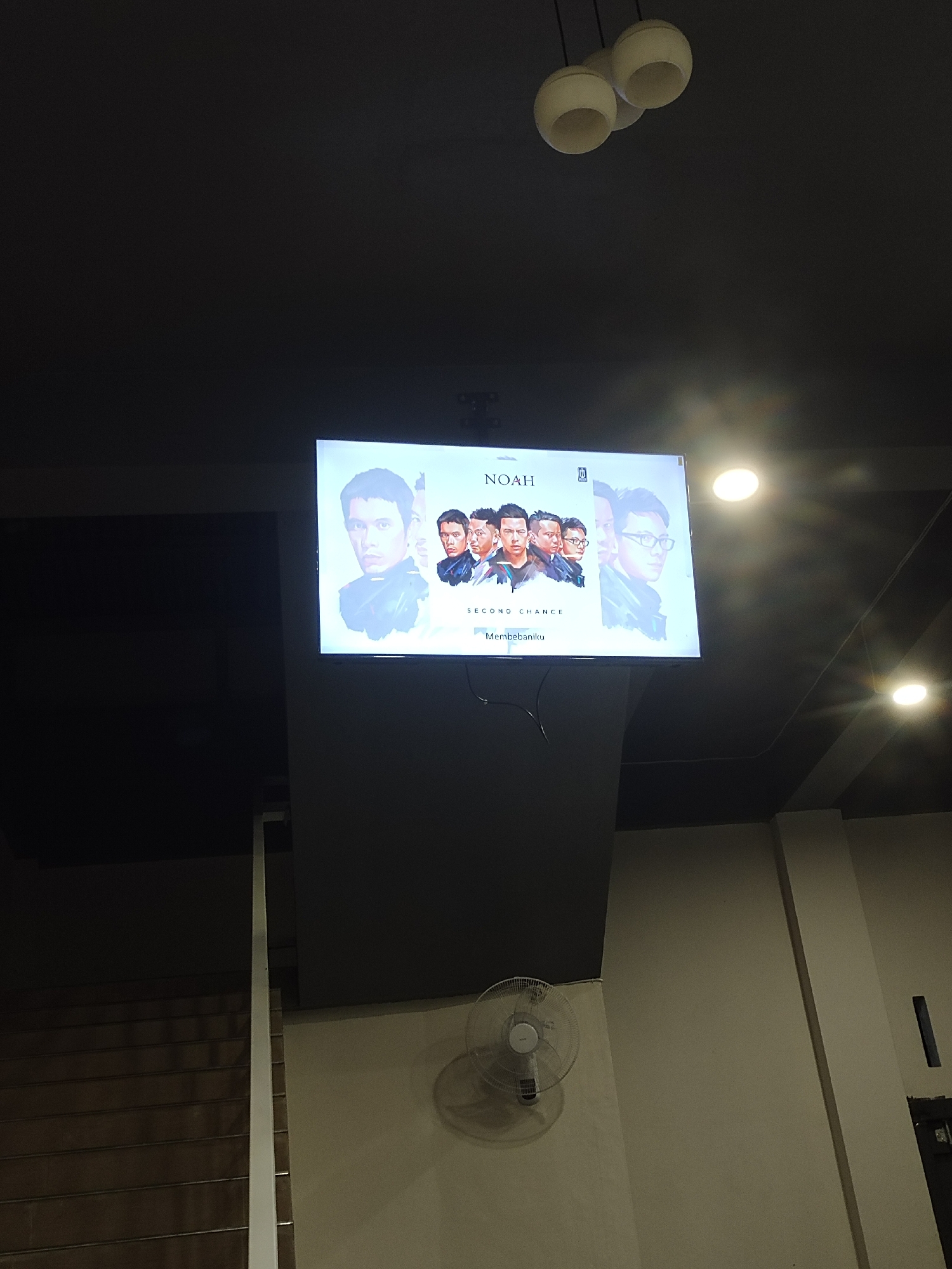 The image depicts the interior of a building in Meunasahaluedrienlhoksukon Barat, Indonesia. A dark grey ceiling is illuminated by three white spherical pendant lights and two bright circular recessed lights. Centrally, a flat-screen television is mounted on a dark grey wall or support structure, displaying an image of the musical group "NOAH" with text "SECOND CHANCE" and "Membebaniku." The television screen shows stylized portraits of five men. To the bottom-right, a white wall-mounted oscillating fan is affixed to a light beige wall, casting a shadow. On the far left, a portion of a brown staircase with a white vertical handrail or wall edge is visible. A dark vertical rectangular object is attached to a wall on the far right side of the frame. The overall lighting is dim, with light sources primarily from the ceiling and the television screen.