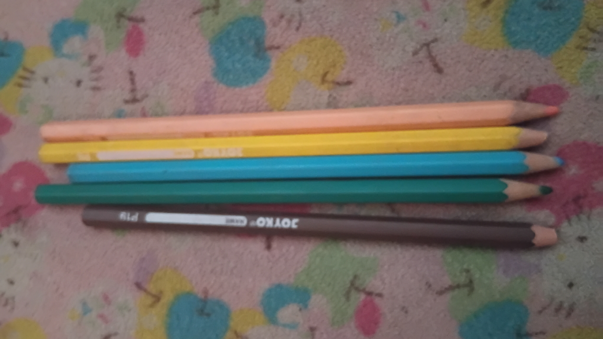 A close-up, overhead shot shows five colored pencils lying on a pink fabric with a cartoon pattern of flowers and animals. The pencils are arranged vertically in a slightly overlapping row from left to right: a brown pencil, a dark green pencil, a light blue pencil, a yellow pencil, and a peach-colored pencil. The brown pencil has white lettering that reads "JOYKO P19". The pencils' tips are pointed, and they are all sharpened. The fabric background is soft and textured, with a repeating pattern of stylized, colorful cartoon figures. The lighting is even, suggesting an indoor setting. There are no people visible in the image. The scene is still and lacks any dynamic action. The image does not provide any information related to the city of Prabumulih, Indonesia.
