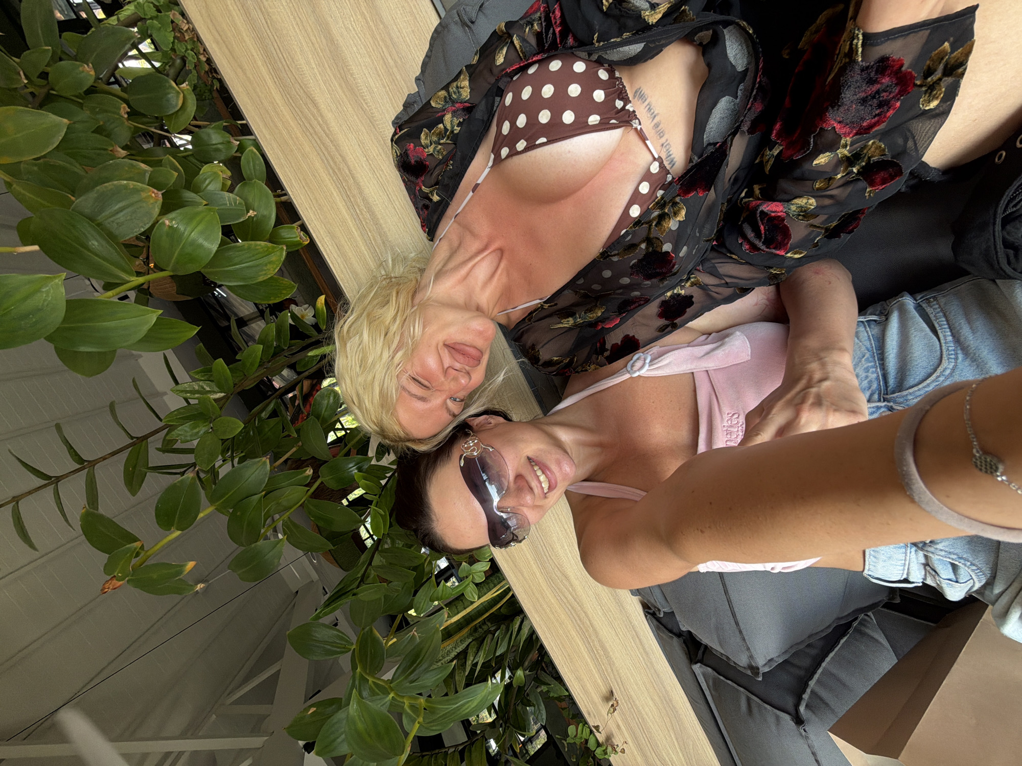 Two women are taking a selfie on a couch. The woman on the left is wearing sunglasses and a pink top. The woman on the right is wearing a brown polka dot bikini top and a black floral robe. They are outdoors, surrounded by lush greenery. The couch appears to be made of dark fabric.