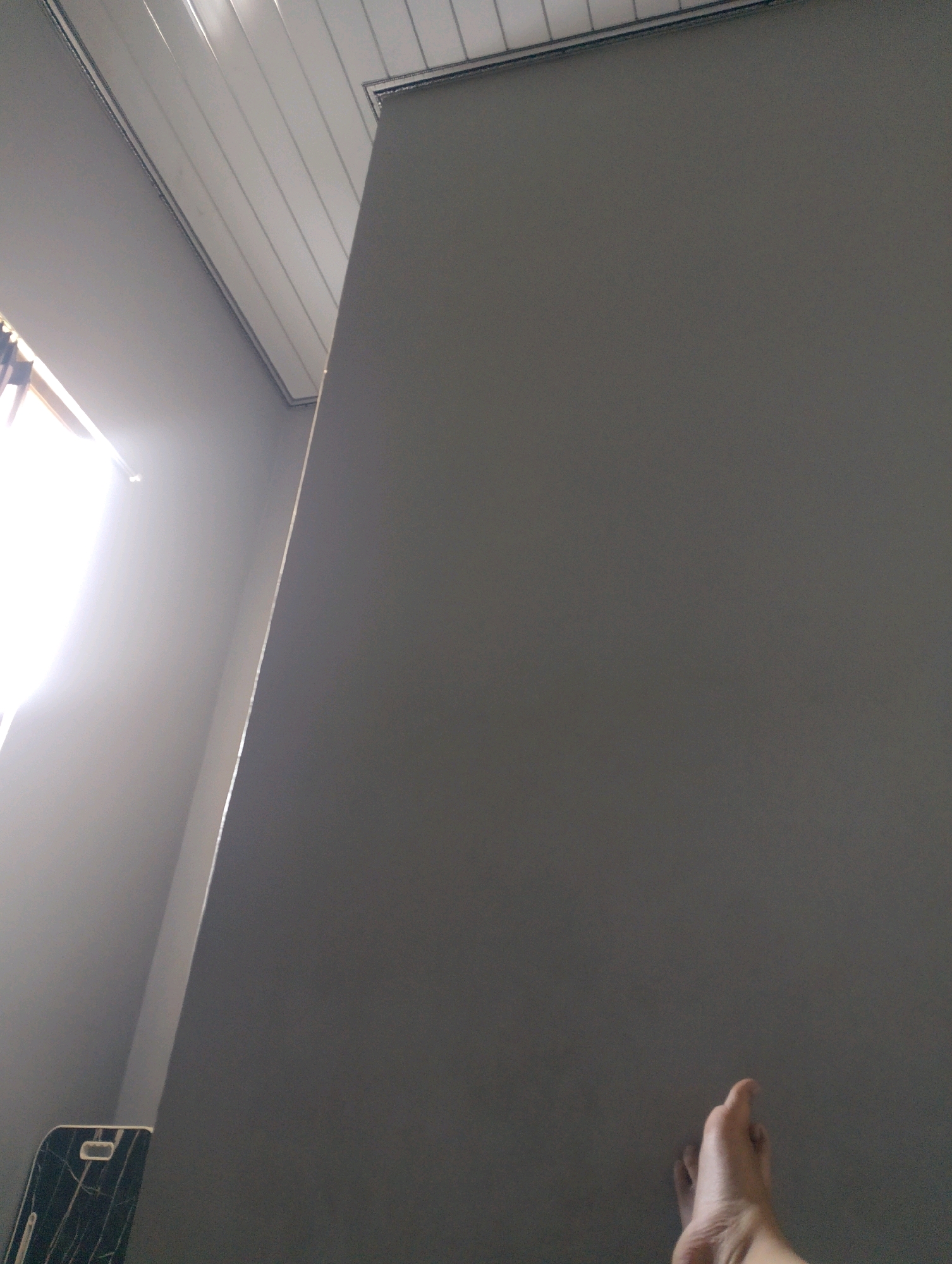 The image depicts an interior scene, likely a room, with a strong light source coming from a window on the left. A human foot is visible in the lower right corner, with the toes pointing upwards. The environment appears to be sparsely furnished, with a large, plain gray wall dominating the right side of the frame. The ceiling, made of white paneling, is visible in the upper portion of the image. A dark, marbled object, possibly a phone or a small appliance, is partially visible in the lower left corner. The lighting creates a dramatic contrast, with the left side of the image being brightly illuminated and the right side in shadow. There is no discernible evidence to place this scene specifically within Magelang, Indonesia, or any other specific city. The image focuses on the interplay of light and shadow, the texture of the wall, and the human foot.
