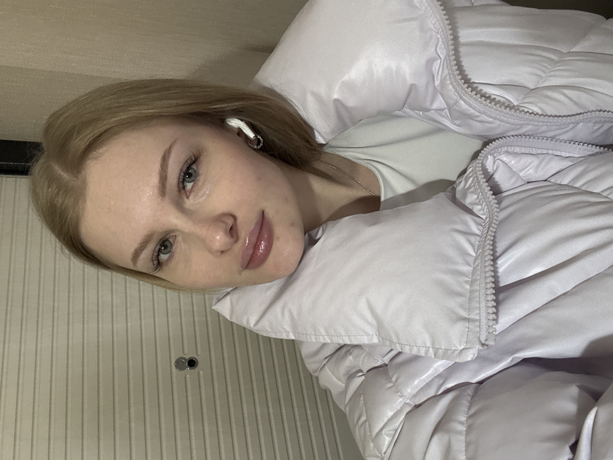A blonde-haired woman wearing a white t-shirt and a lavender puffer jacket is positioned in the center of the frame, facing the camera. An earbud is visible in her left ear. Behind her, the background includes a door. The location is identified as Perm, Russia.