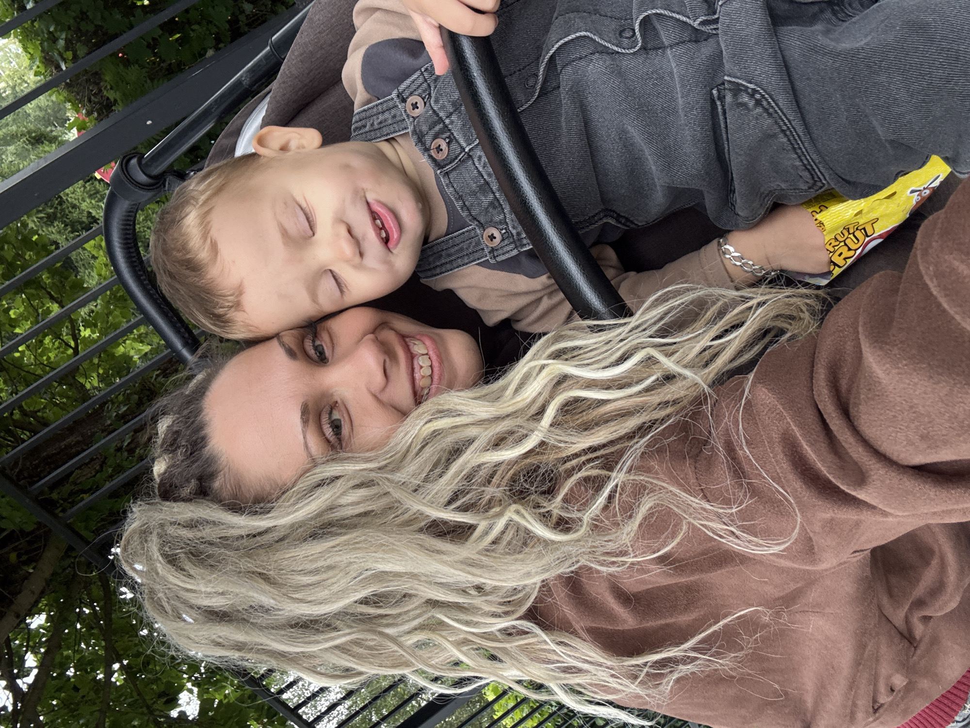 In the city of Rusanovo, Russia, a woman with blonde, heavily highlighted hair pulled into a high ponytail and a brown hooded jacket is taking a selfie with a young child. The child, wearing denim overalls over a brown long-sleeved shirt, is seated in a stroller, with their eyes closed and a slight smile on their face. A bright yellow candy wrapper is visible tucked into the stroller beside the child. The background shows a black metal fence and green foliage, suggesting an outdoor setting. The interaction captured is a moment of shared affection and casual outdoor activity.