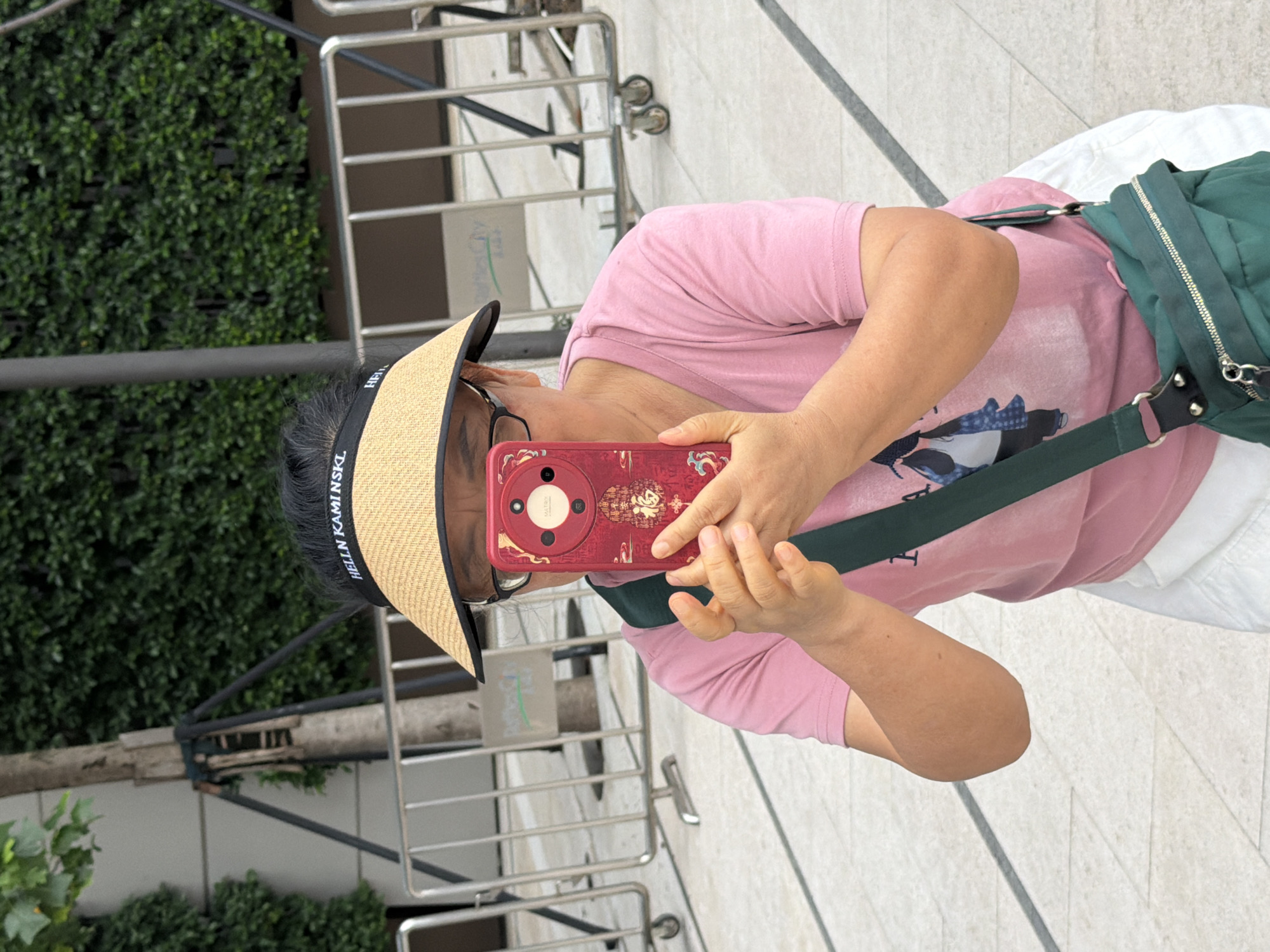 A woman in a pink t-shirt, white shorts, and a wide-brimmed straw hat is holding a red smartphone in front of her face, appearing to take a photo or video. The phone case has an intricate design with red and gold accents, and possibly a Chinese character that translates to "fortune". She wears glasses and has dark hair pulled back. A dark green crossbody bag is slung over her right shoulder. The background shows a paved walkway with diagonal grey lines, metal barricades, a tree, and lush green foliage. The scene appears to be outdoors in an urban environment, potentially in the city of Yuzhong District, China, given the style of the phone case and the presence of urban elements.