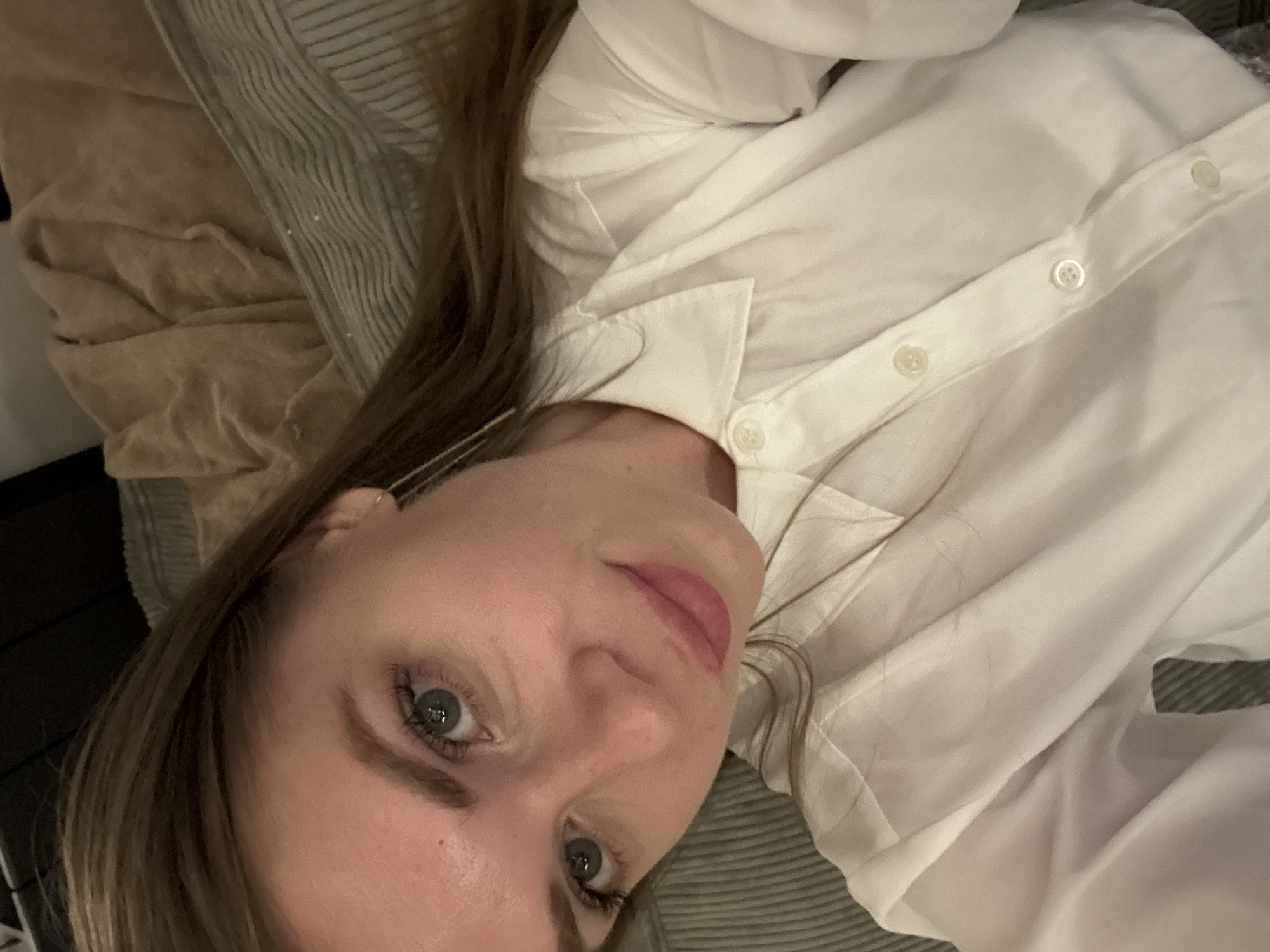 A woman with light skin and gray eyes is the central subject of the image. She has light brown hair. She is wearing a white button-down shirt. She is lying down on a striped surface. A tan colored, wrinkled fabric is behind her left side. The photograph does not reveal a location in Batumi, Georgia.