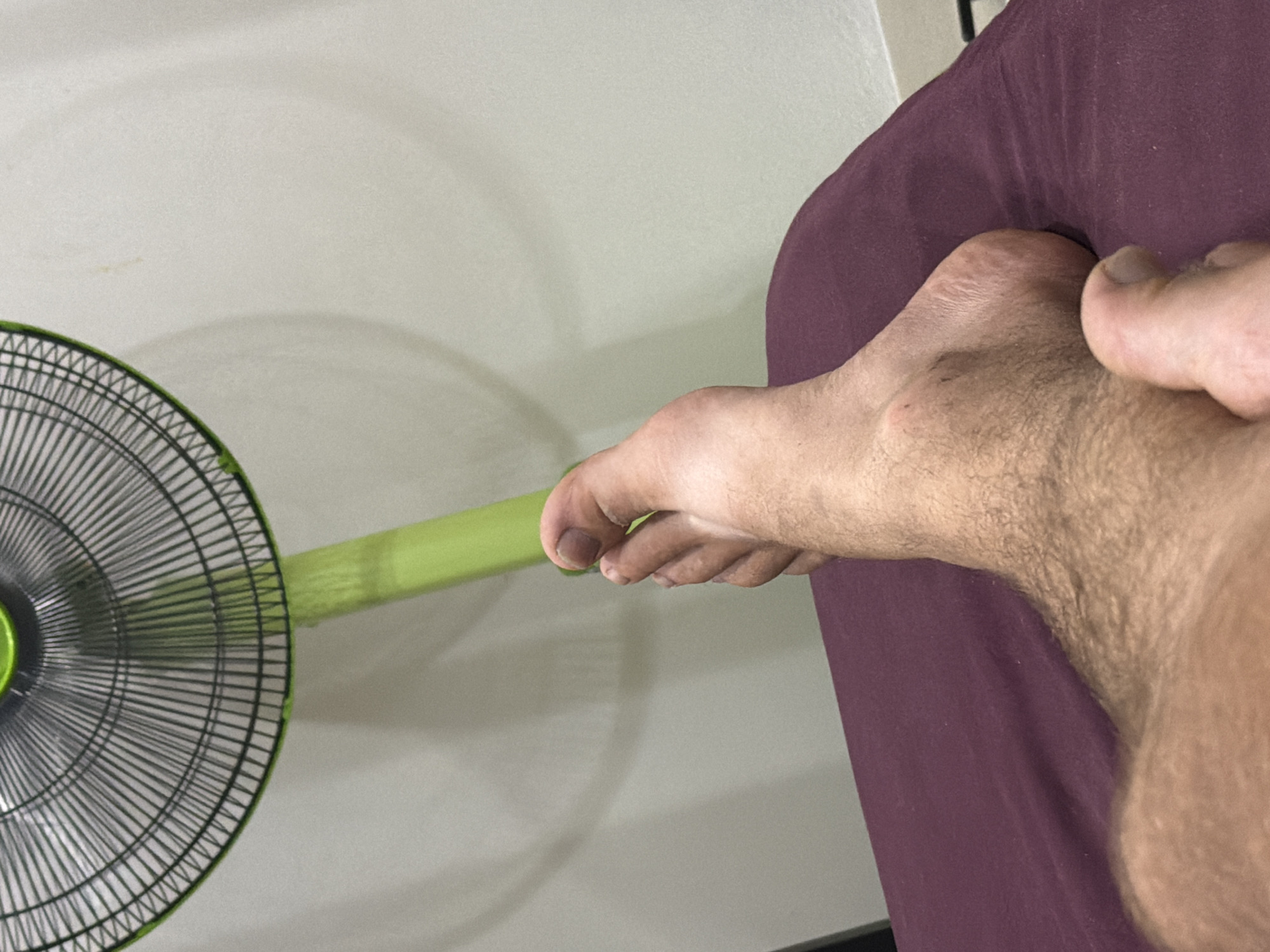 A person's bare foot is resting on a plum-colored surface, with a portion of their leg visible. Behind the foot and leg, a green fan is positioned, casting a shadow on a white wall. The fan has a green frame and a black grate. The scene appears to be indoors, possibly in a bedroom or living area. The overall lighting is soft, suggesting ambient room light. No specific location in Baan Mae Nam, Thailand, is identifiable from the image.