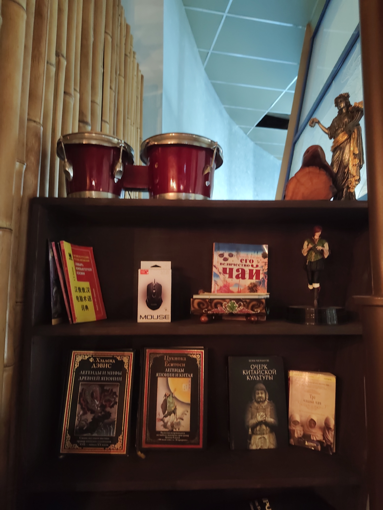 This is a medium shot, indoor, eye-level, horizontal photograph of a dark wooden shelf filled with books and decorative items. The shelf is the main subject of the image.
On the top shelf, two red bongo drums are prominently displayed. Behind them, a wall covered in vertical bamboo stalks is visible. To the right of the drums, a golden statue of a woman holding flowers stands next to a reddish-brown, abstract sculpture. The background on the top shelf is a light blue wall with a white ceiling with visible grid lines.
The middle shelf contains several items. On the left, three books are leaning against each other. The front book is red with yellow text and what appears to be Chinese characters. To its right, a white box labeled "MOUSE" with a black computer mouse image is positioned. In the center of the shelf, a colorful book titled "его невероятное чаи" (its incredible tea) sits on a decorative wooden stand with floral motifs. To the right of the book, a figurine of a person in historical clothing stands on a dark circular base.
The bottom shelf is filled with more books. On the left, a book with a black cover and gold lettering titled "Ф. Хэллена Дэвис Легенды и мифы древней Японии" (F. Hellena Davis Legends and myths of ancient Japan) is visible. Next to it, a book with a similar style of gold lettering and a picture of a woman in traditional clothing is titled "Лукноса Еситоси Легенды Японии и Китая" (Luknosa Yoshitoshi Legends of Japan and China). Further to the right, a dark green book with gold text reads "Очерк китайской культуры" (Essay on Chinese culture) and features an image of a statue. To the right of that, a light-colored book with the title "Три чашки чая" (Three cups of tea) is displayed.
The lighting in the image is somewhat dim, casting shadows on the shelf and its contents. The overall environment appears to be an indoor display, possibly in a shop or a private collection.
The city of Sochi, Russia is not mentioned or implied in the image or its content.
