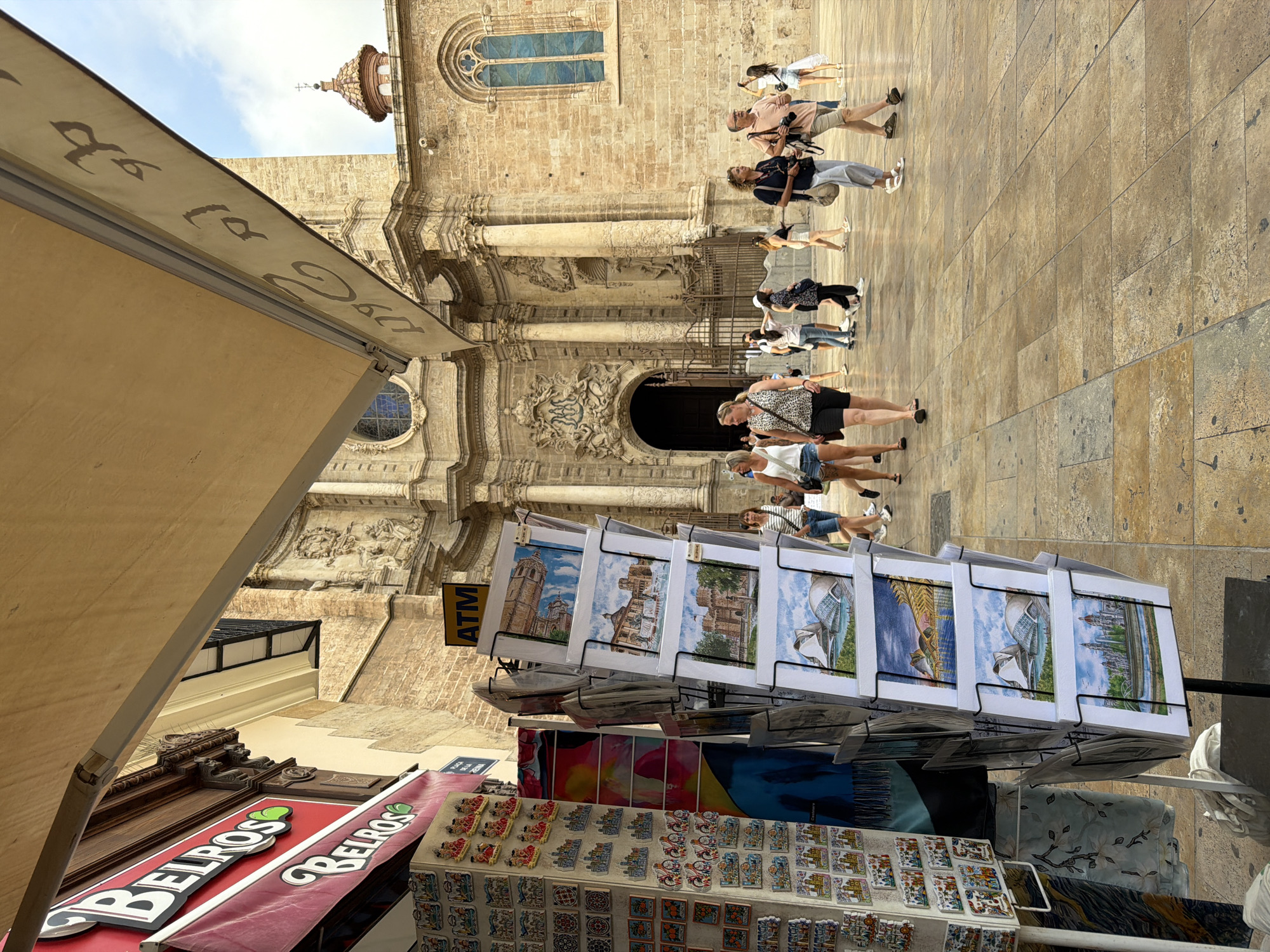 A postcard stand displays various scenic cards in front of an ornate stone building, likely a cathedral, with a tall bell tower and a dome visible. The building's facade features intricate carvings and a large arched entrance. Several people are walking across the paved plaza in front of the building. The postcard stand exhibits images of local landmarks, including what appears to be the City of Arts and Sciences in Valencia. The scene is illuminated by natural daylight, suggesting it is daytime. A red awning with the store name "BELROS" is visible on the left. An ATM sign is also mounted on the building. The overall environment is an urban public space.