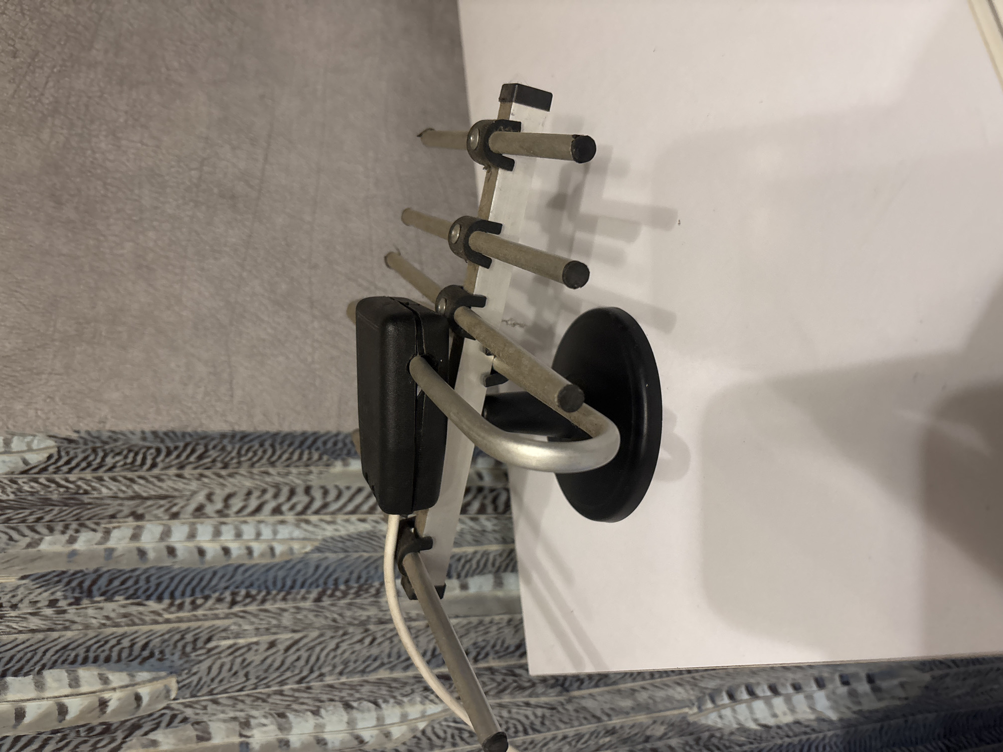 A tabletop indoor television antenna sits on a white surface, possibly a table or desk. The antenna has a black rectangular base and a silver, curved stand. It features several silver rods extending from a central silver bar, designed to receive broadcast signals. A white cable is attached to the antenna. In the background, a textured grey wall is visible, and to the left, a decorative hanging object with a pattern resembling feathers is present. The lighting suggests an indoor setting.