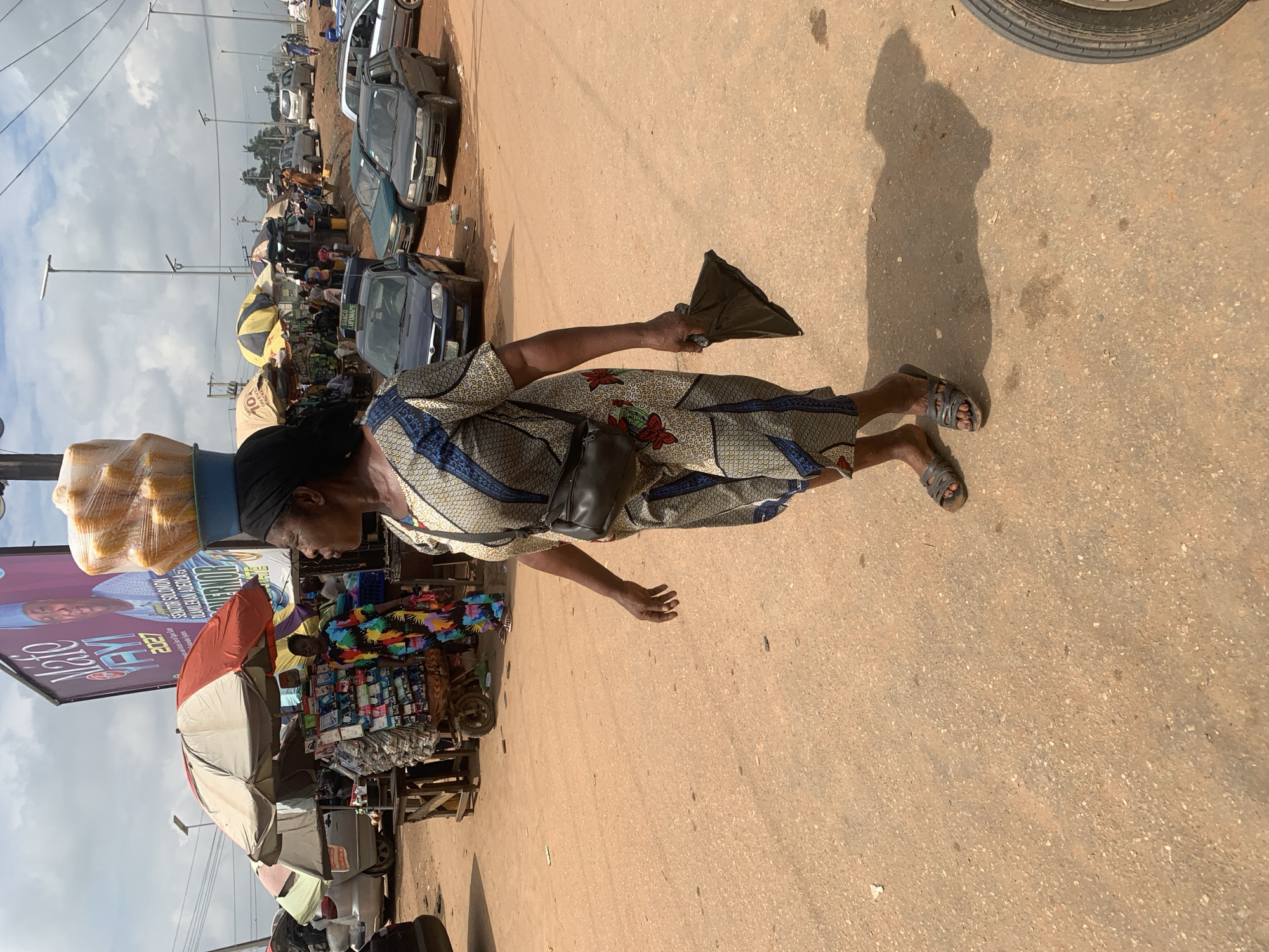 In Shagamu, Nigeria, on a dusty, unpaved road, a woman walks past a market stall. She carries a large basket of corn on her head and wears a patterned dress and sandals. Behind her, market stalls are set up under umbrellas, and cars are parked on the side of the road. A large banner with a political advertisement is visible in the background. The scene is bustling with activity, typical of a Nigerian market on a bright, sunny day.
