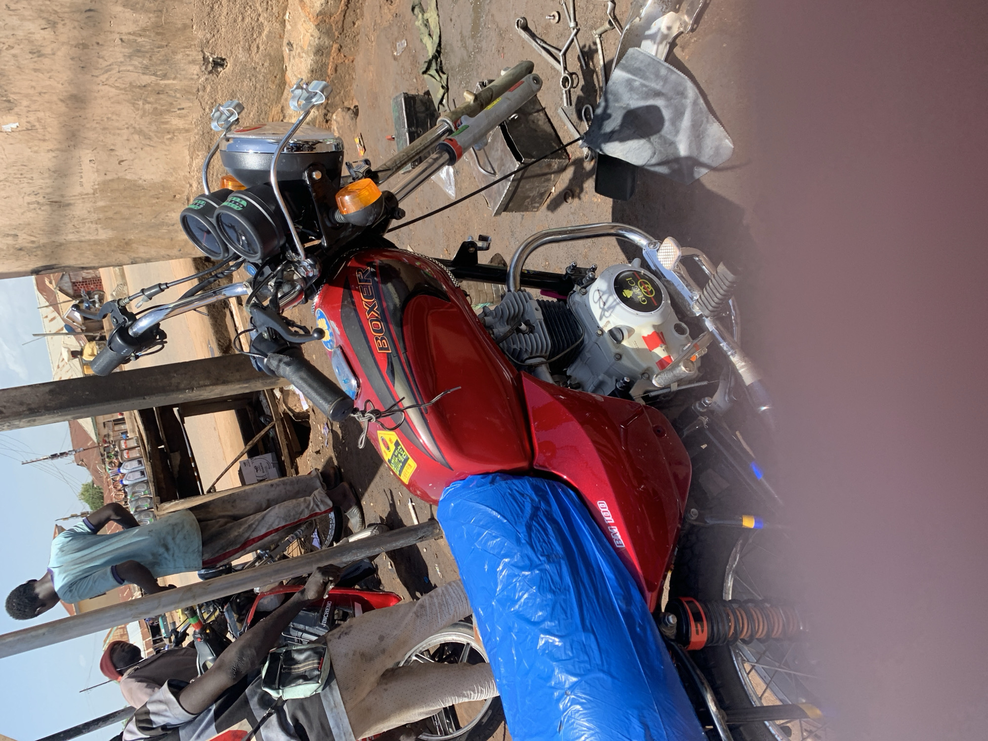 The image, rotated 90 degrees clockwise, depicts a red Bajaj Boxer motorcycle parked outdoors in what appears to be a informal workshop or roadside area in Gura Top, Nigeria. The motorcycle is the primary subject, dominating the frame. Its fuel tank is a vibrant red with "BOXER" emblazoned in black and orange, and "BAJ 100" visible on its side. A bright blue plastic sheet covers the seat and extends down, obscuring parts of the frame.
The front of the motorcycle shows its handlebars, chrome mirrors, and a dashboard with two circular gauges. An orange turn signal is visible on the right. The engine, with its cooling fins, has a white cover adorned with a yellow and green "Gucci" logo sticker. Various tools and metal parts are scattered on the dirt ground surrounding the motorcycle, suggesting a repair or maintenance setting.
In the background, two men are visible. One man, wearing a light blue t-shirt and light-colored trousers, stands near the motorcycle. Another man is partially visible, seated in the lower left portion of the image, next to what appears to be another motorcycle or bicycle frame with "JINCHENG" visible on it. The scene is bathed in bright daylight, indicated by strong shadows. The general surroundings include simple structures and hints of other vehicles in the distance, typical of a bustling street or roadside in the region. No specific activity is captured beyond the presence of the motorcycle and the individuals.