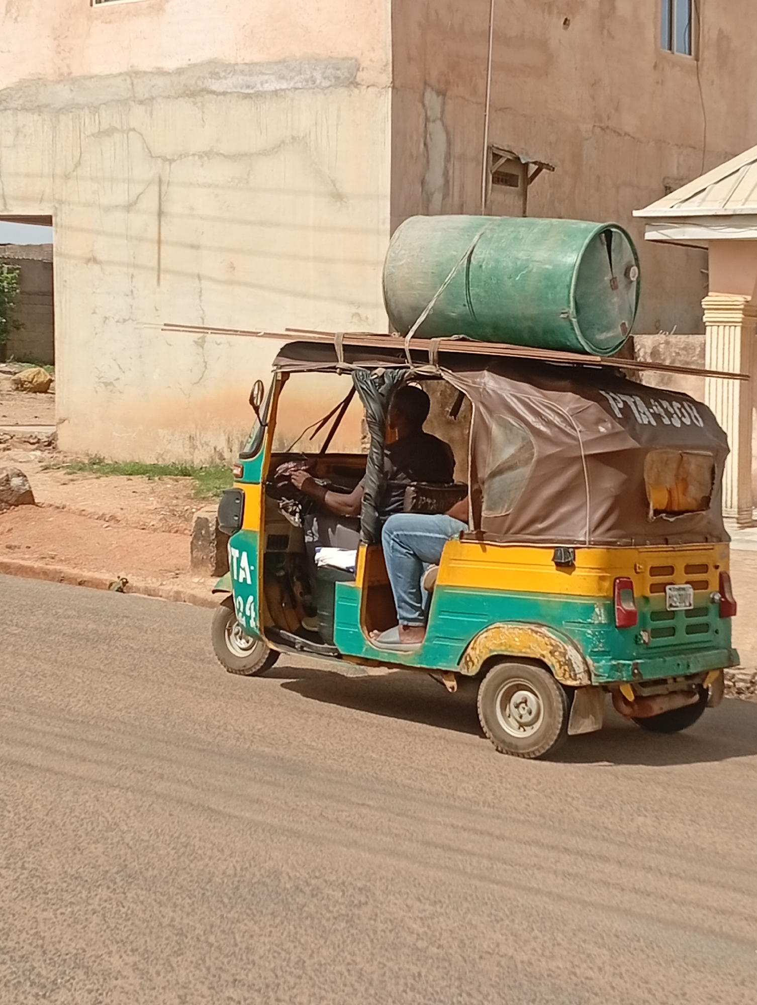 The image depicts a three-wheeled vehicle, a "keke," driving on a paved road in Barakin Akawo, Nigeria. The keke is painted in green and yellow and has a brown canvas roof. A man is driving the keke, wearing a black shirt and blue jeans. On the roof of the keke is a large green plastic barrel secured with metal bars. The license plate of the keke, "PTA-4330B," is visible on the roof covering. Buildings are visible in the background. The overall scene appears to be a regular street scene in a residential or commercial area of Barakin Akawo.