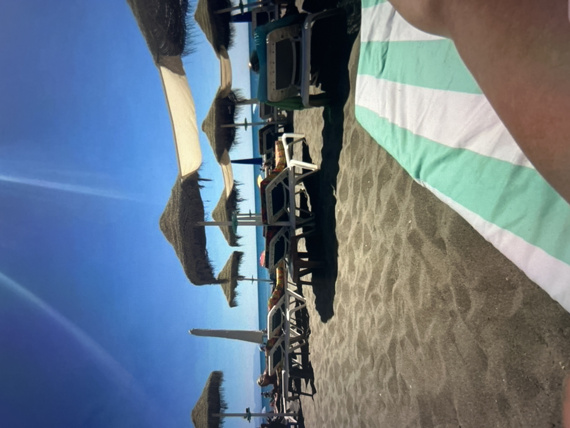The image depicts a sunny beach scene, viewed from a first-person perspective, likely from a beach chair. In the foreground, partially visible on the right, is a person's arm and a light green and white striped towel spread on dark, possibly volcanic sand.
The midground features several rows of beach umbrellas and sun loungers stretching across the dark sand. Many of the umbrellas are made of straw or thatch, while others are made of light-colored fabric. Some sun loungers appear empty, while a few have people relaxing on them, though their figures are mostly obscured or distant. The sand shows ripples and texture, indicating footprints or wind patterns.
In the background, a serene, light blue sea meets a clear, vibrant blue sky. A prominent diagonal light streak or lens flare is visible in the upper left portion of the sky, suggesting the sun's position.
The scene conveys a sense of a leisurely day at a beach resort, characterized by bright daylight and fine weather. There is no discernible text visible in the image.