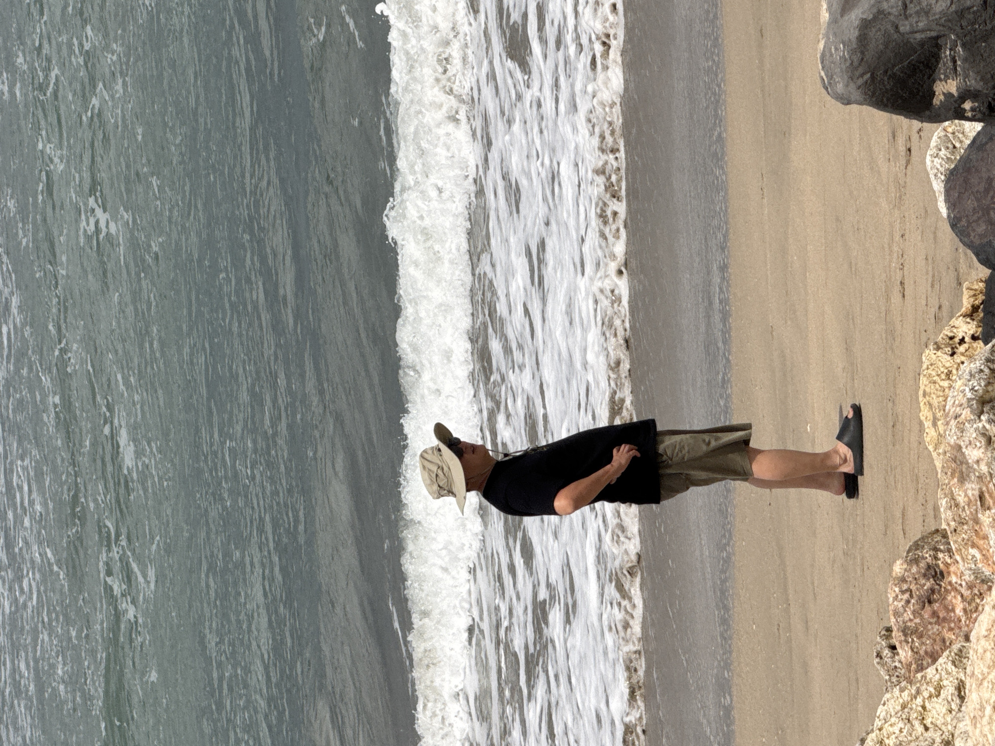 An adult male, wearing a black t-shirt, khaki shorts, a wide-brimmed hat, sunglasses, and black slides, stands on a sandy beach in Kuta, Indonesia. He is facing right, with his right hand on his hip. The ocean is behind him, with waves breaking and white foam visible. Rocks are in the foreground.