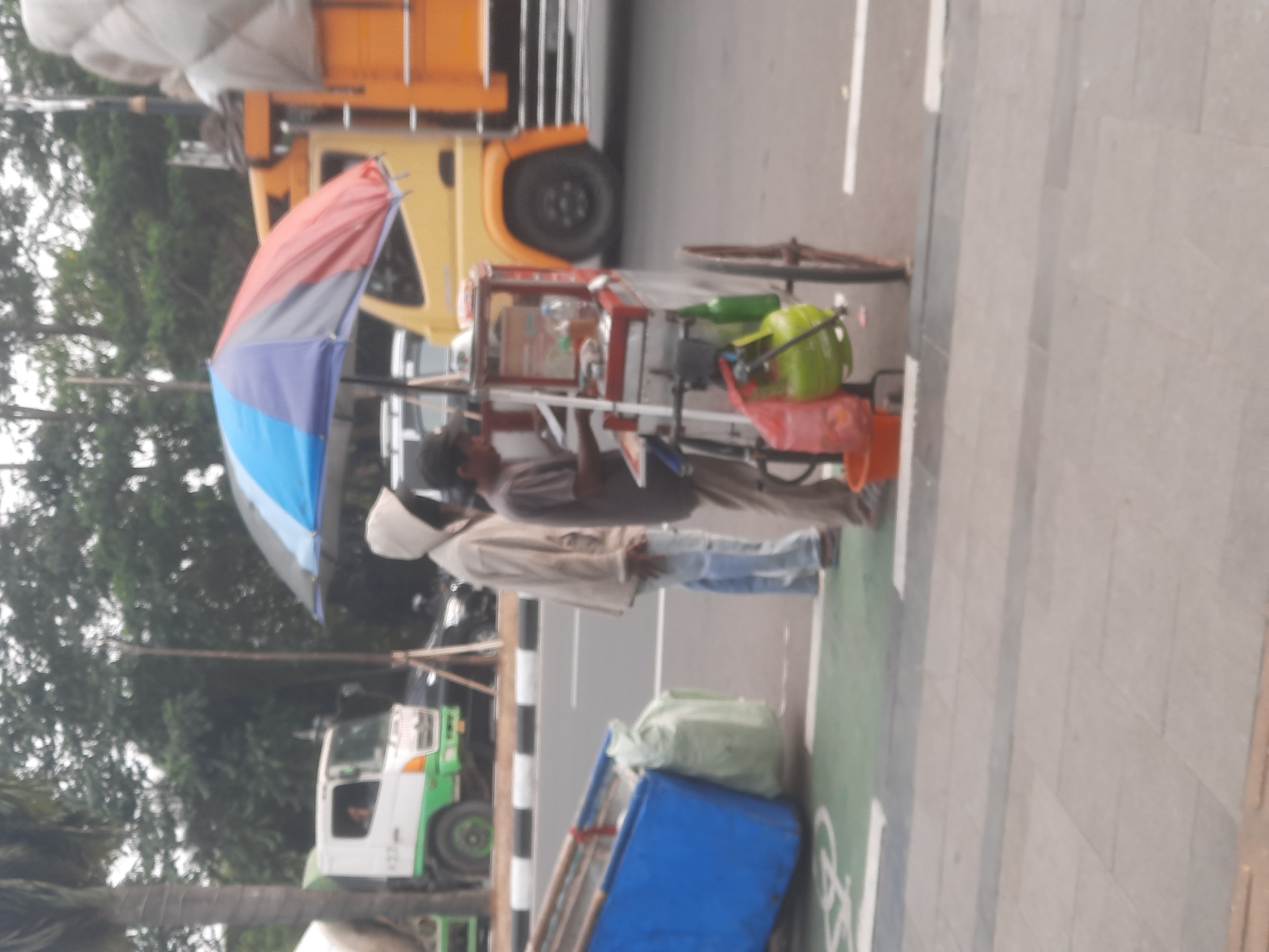 The image depicts an outdoor scene, possibly in Sukaraja, Indonesia, on a street with a sidewalk and a bike lane. In the foreground, a street food cart is visible, equipped with a propane tank and various containers. Two individuals are present; one, wearing a hooded jacket and jeans, stands near the cart, while another, in a t-shirt and pants, appears to be operating or attending to the cart. Behind them, a yellow truck with an orange bed is parked on the road. A white cement truck is also present in the background. The environment suggests a typical urban street setting with trees lining the area.