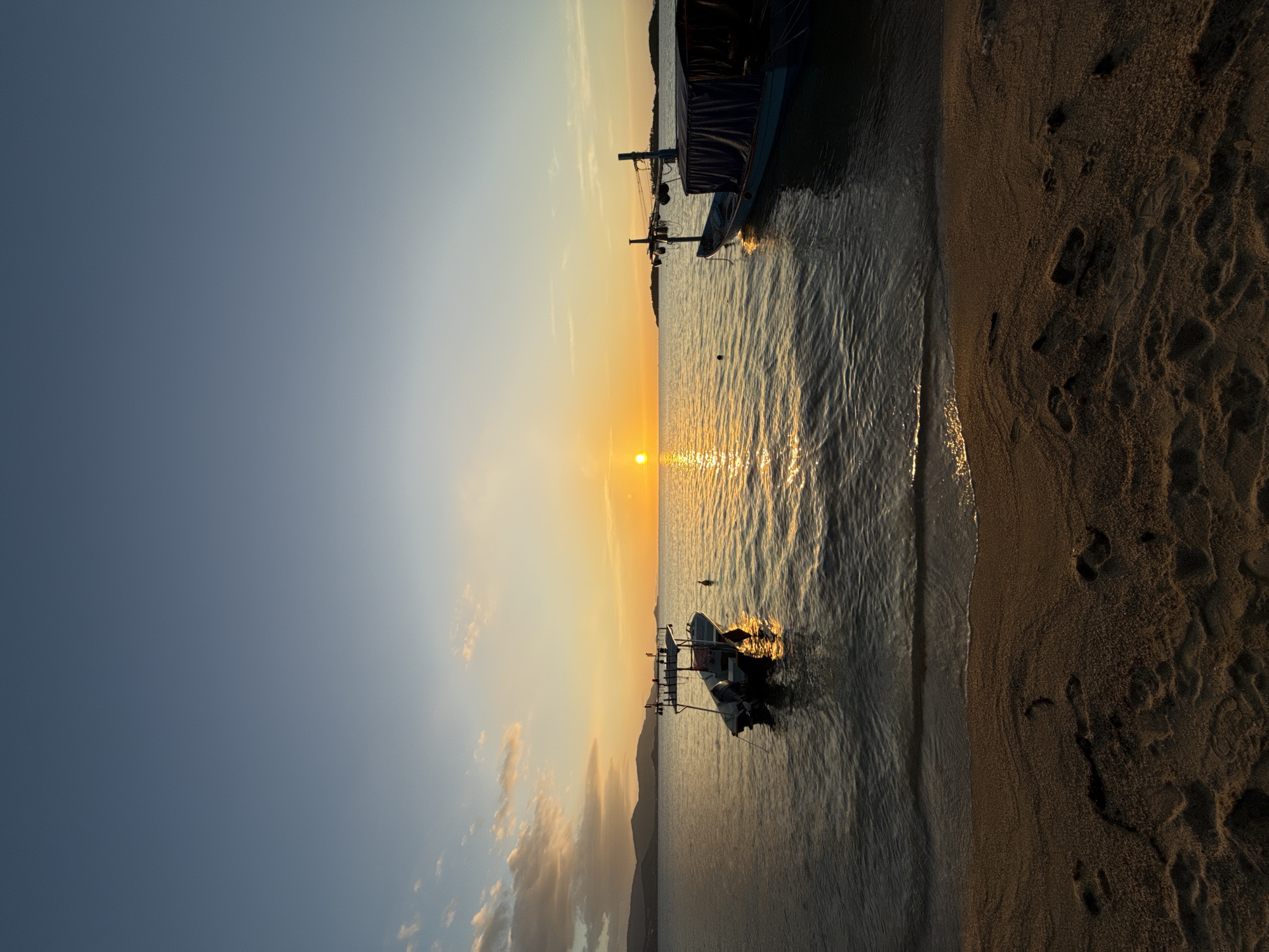 A serene sunset over the Gulf of Thailand, viewed from the shore in Ko Samui District, Thailand. The sun, a brilliant orb, hovers just above the horizon, casting a golden glow across the tranquil water. Gentle waves lap at a sandy beach in the foreground, where footprints are visible, suggesting recent human presence. Two boats are anchored offshore: a smaller, modern vessel with a canopy in the left mid-ground, and a larger, traditional fishing boat with a covered section to the right. Distant hills form a silhouetted backdrop against the vibrant sky, painted with streaks of orange, pink, and pale blue. The scene is peaceful, with no people immediately visible.