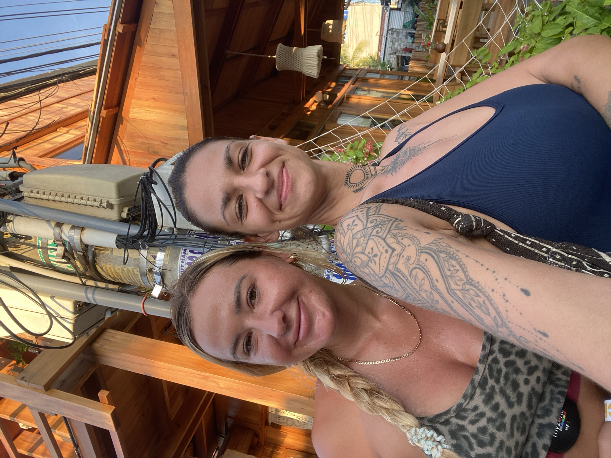 Two women are posing for a selfie in an outdoor setting. The woman on the left has blonde hair in a braid and is wearing a leopard-print top and a gold chain. The woman on the right has dark hair pulled back and is wearing a dark blue tank top. Both women have tattoos. They are standing in front of a wooden structure with a thatched roof, possibly part of a restaurant or bar. In the background, there are trees, other buildings, and a street light pole with many wires. The lighting suggests it is late afternoon or early evening. The scene appears to be located in Mũi Né, Vietnam, based on the architectural style and vegetation.