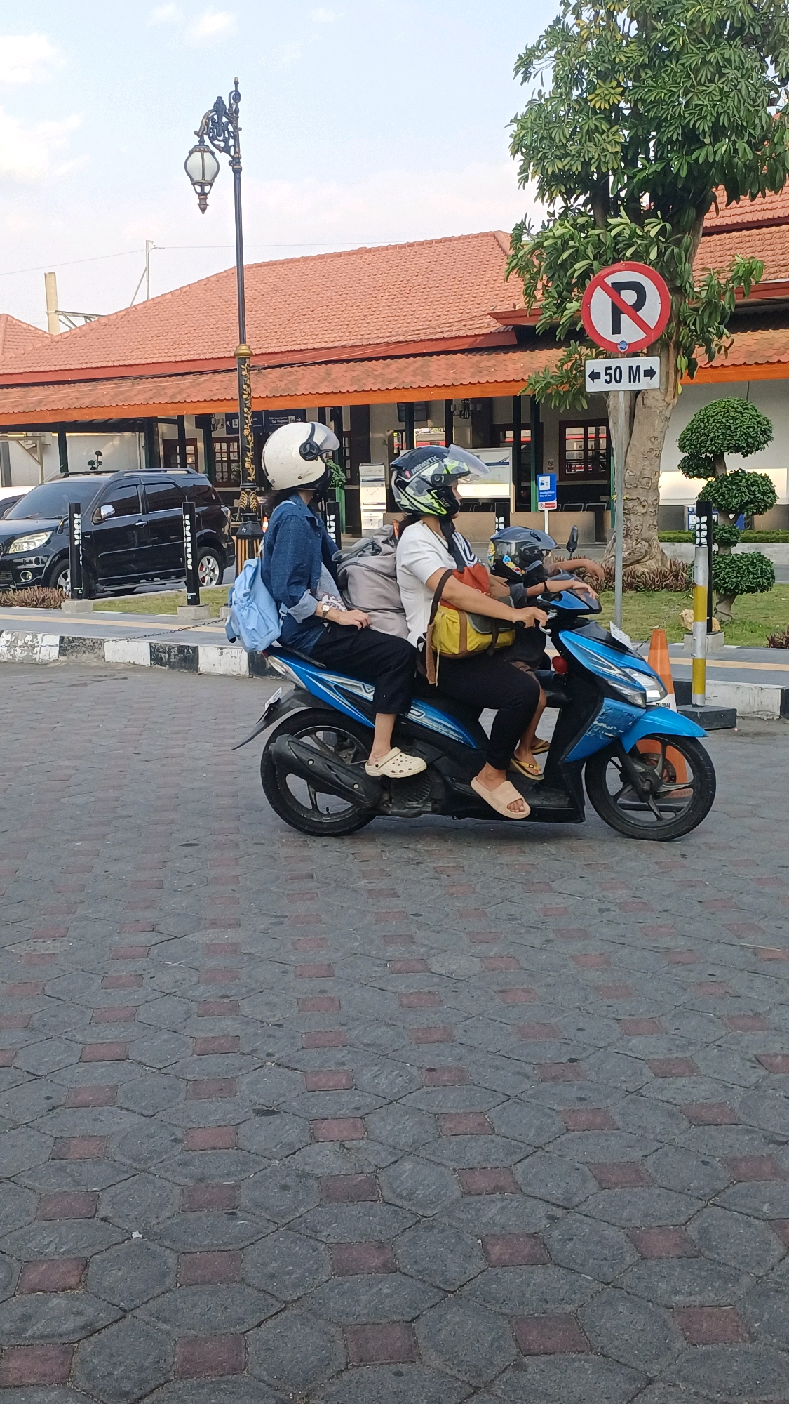 The image shows two people on a blue scooter in front of a building in Klaten, Indonesia. The scooter is moving from left to right. The person in the back is driving, and the person in the front is a passenger. Both people are wearing helmets. The building behind them appears to be a train station, with a tiled roof and a decorative lamppost. There is a "no parking" sign with an arrow pointing to the right, indicating 50 meters. A black SUV is parked on the left side of the image. The ground is paved with interlocking stones. The sky is partly cloudy.