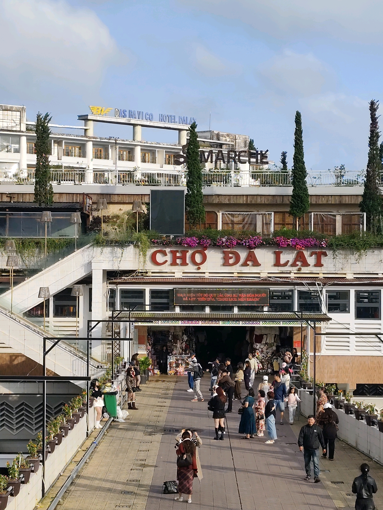 The image depicts a bustling marketplace scene in Da Lat, Vietnam, likely in the vicinity of the Da Lat Market, known for its vibrant atmosphere and diverse offerings. In the background, the prominent "DASHAVI CO HOTEL DALAT" signage is visible, indicating a hotel establishment. Further to the right, the word "MARCHE" is also displayed, suggesting a market or commercial area.
The central focus of the image is the "CHO DA LAT" sign, a clear indication of the Da Lat Market. Below this sign, numerous stalls are set up, showcasing various goods. People are seen actively engaged in shopping and strolling through the market. A diverse crowd of individuals, including men, women, and children, are navigating the crowded pathways. Some are examining merchandise, while others are captured in candid moments of movement and interaction.
The architecture of the buildings suggests a multi-level structure with balconies and open-air walkways. Planters filled with green foliage and colorful flowers adorn the balconies and walkways, adding a touch of natural beauty to the urban landscape. The ground beneath the people is paved, with distinct lines suggesting paving stones. The sky above is partly cloudy, with a soft, diffused light illuminating the scene. Several tall, slender trees are strategically placed throughout the area, contributing to the overall aesthetic. The presence of stairs and railings indicates an accessible and organized layout for pedestrians.