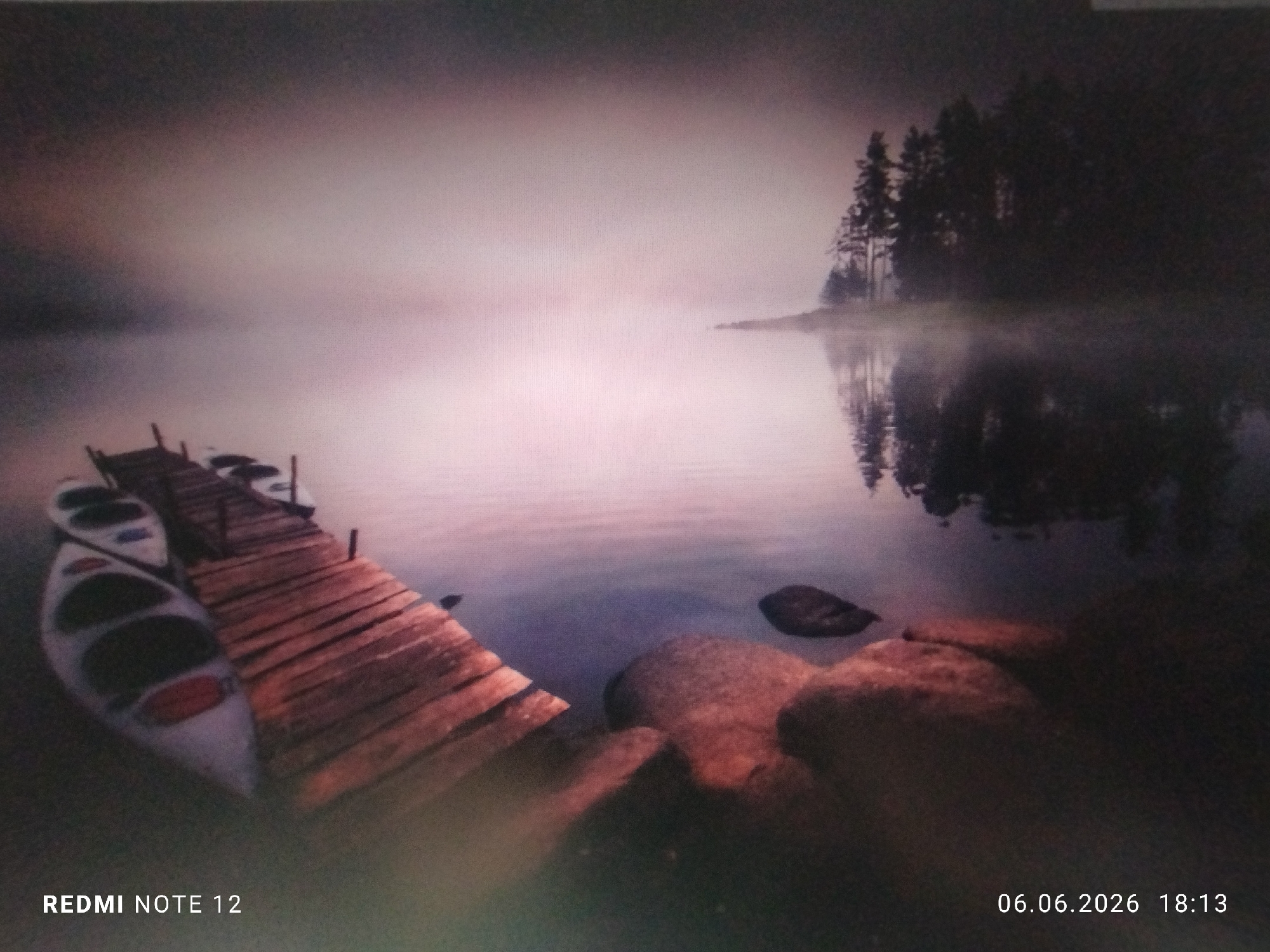 The image displays a serene natural landscape at dusk or dawn, characterized by a thick mist hovering over a calm body of water. A wooden dock, constructed from planks, extends diagonally from the lower-left foreground towards the center. Several small, white canoes or kayaks with red accents are moored along the left side of the dock.
The water surface in the midground is largely obscured by a diffused, pinkish-purple mist that extends across the scene, blending with the sky in the distance. On the right side, a small landmass or island emerges from the mist, populated by a cluster of tall, dark evergreen trees, which are clearly reflected in the still water below the mist layer. In the bottom-right foreground, several large, reddish-brown rocks are partially submerged along the water's edge.
No people are visible in the scene. The environment is tranquil and appears remote, lacking any urban identifiers or landmarks specific to the city of Burgas, Bulgaria. The lighting is soft and diffused, creating an atmospheric, subdued ambiance. Text overlays on the image read "REDMI NOTE 12" in the bottom left and "06.06.2026 18:13" in the bottom right.