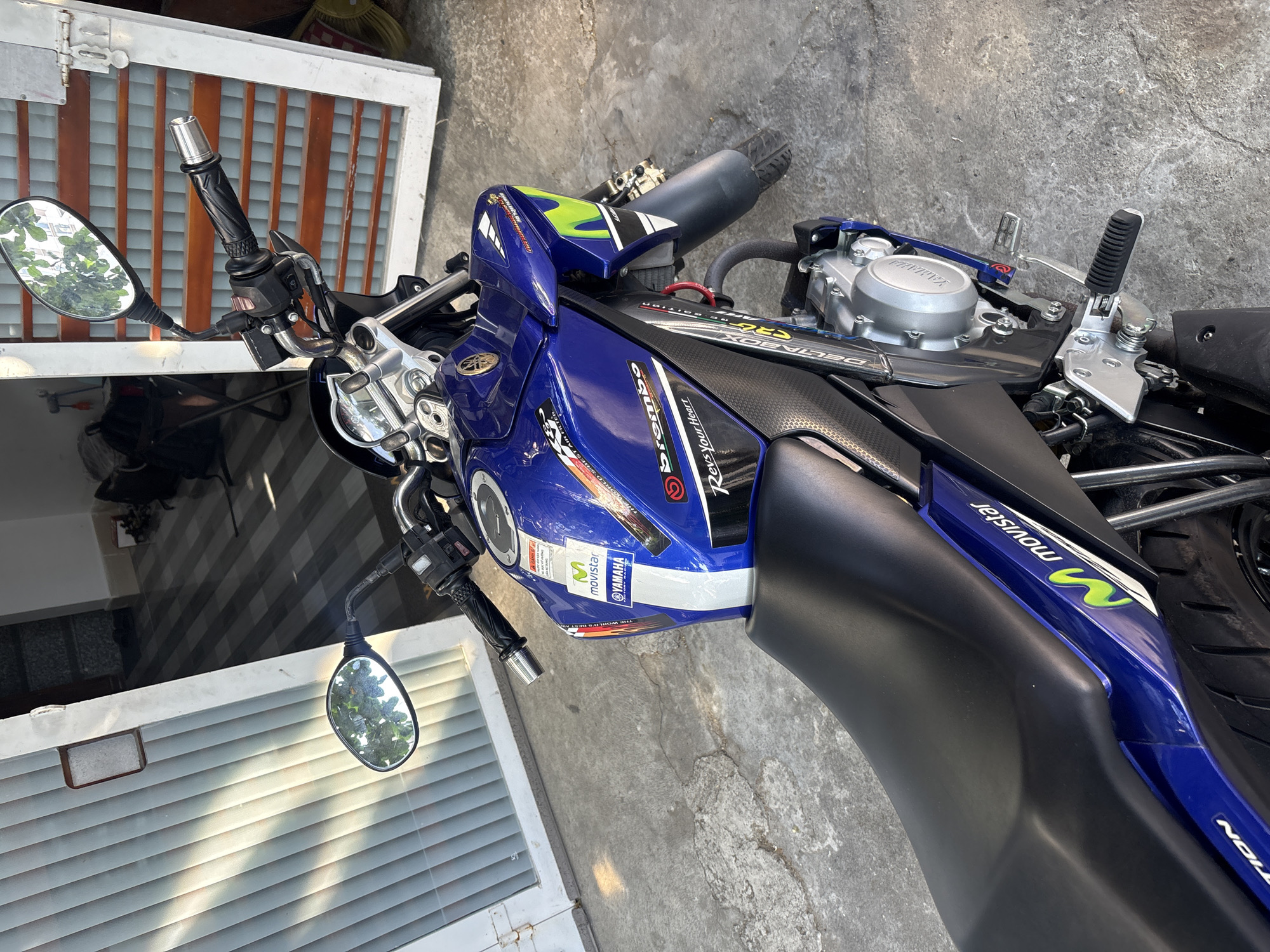 The image shows a top-down view of a blue and black Yamaha motorcycle parked on a concrete surface. The motorcycle has various decals and stickers, including "Brembo," "Movistar Yamaha," and "Yamaha" logos. The engine is visible, with a silver-colored cover. The motorcycle's mirrors are present on both sides. The background includes a white window frame with wooden slats and a glimpse of a shadowed area with plants. The image was taken in the Nha Trang Ward area, Vietnam.