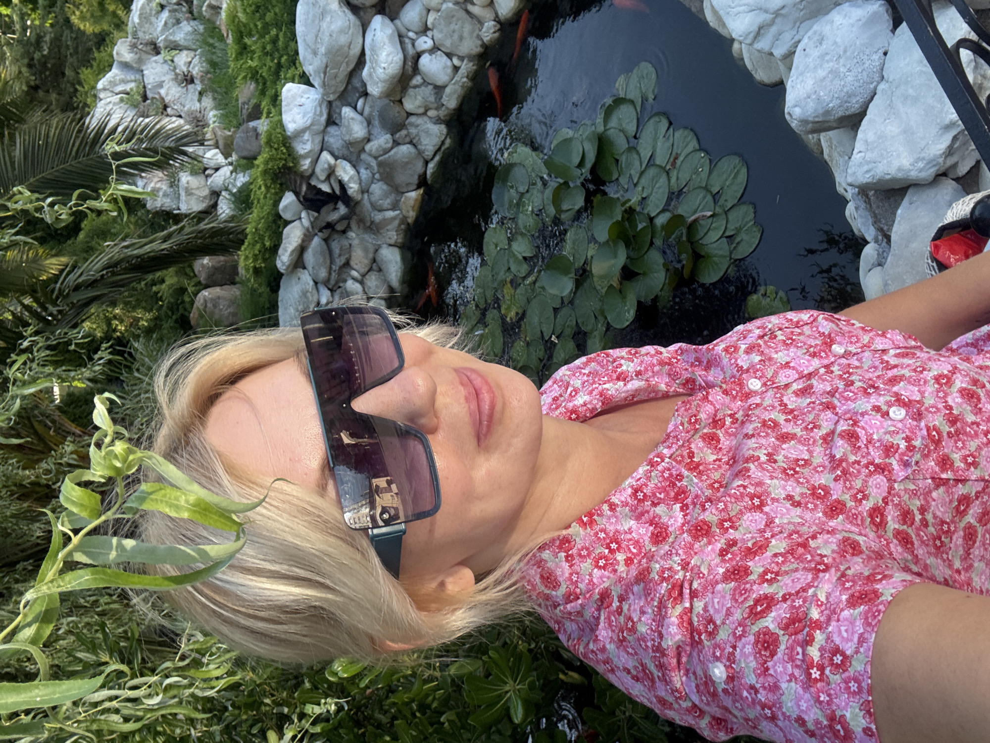 In a sun-drenched outdoor setting, likely within the verdant landscape of Sochi, Russia, a blonde woman with fair skin and a smattering of freckles is captured in a medium shot, looking towards the viewer's right with a slight smile. She is adorned in a short-sleeved pink blouse patterned with a dense floral print of red and white blossoms. Her face is partially obscured by large, dark, angular sunglasses with a teal frame, the lenses reflecting a building with arched windows, suggesting an urban or architectural element nearby. Her blonde hair is styled casually, with strands framing her face.
The background reveals a meticulously crafted garden scene. To the left, lush green foliage, including weeping branches, cascades downwards, suggesting a naturalistic yet manicured environment. Dominating the right side of the frame is a decorative pond, its dark water dotted with numerous green lily pads. Several bright orange koi fish are visible swimming beneath the surface, adding a vibrant splash of color. The pond is bordered by a wall constructed of varied sizes of smooth, light-colored stones. Behind the pond, a rocky outcrop, also adorned with stones and interspersed with green vegetation, rises, creating a naturalistic backdrop. Further in the distance, palm trees and other diverse greenery indicate a subtropical climate, consistent with Sochi. The overall impression is one of a serene and picturesque garden, possibly a public park or a private estate.