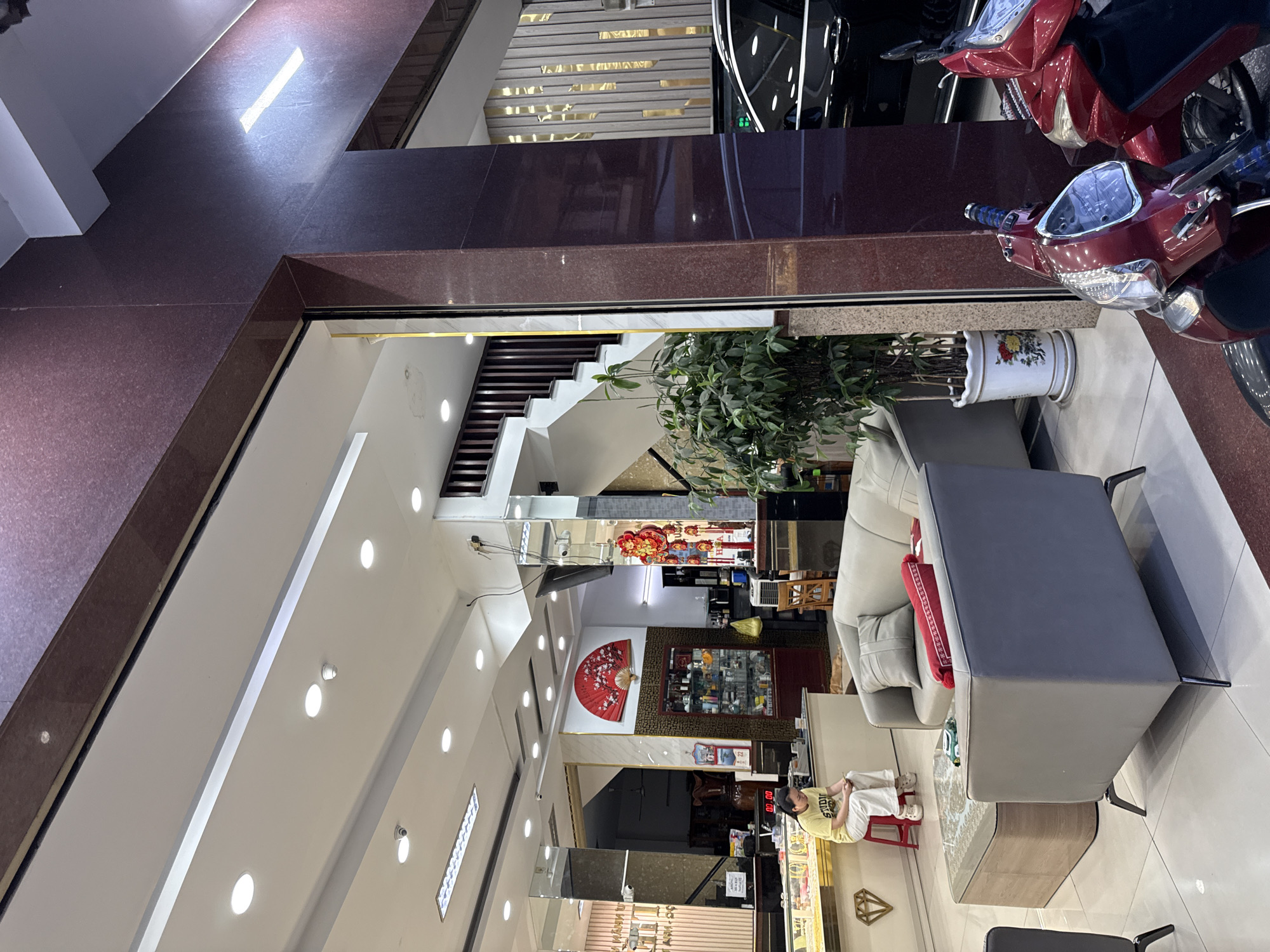 The image depicts the interior of what appears to be a jewelry store or a high-end retail establishment in Nha Trang, Vietnam. The environment is modern and well-lit, with a combination of recessed lighting and strip lights. In the foreground, a woman in a yellow t-shirt and white pants is seated on a red stool, looking down at her phone. She appears to be a customer or employee. To her left is a counter displaying jewelry. In the center of the frame, a gray sectional sofa with a red and gray accent pillow is arranged around a glass-topped coffee table. To the right of the sofa, a large potted plant with dark green leaves adds a touch of nature to the scene. Behind the plant, a dark-colored car is visible through an open doorway. Red motorbikes are parked just outside the entrance to the establishment. The walls are decorated with a large red fan motif and display shelves stocked with various items. A staircase is visible in the background, suggesting multiple levels within the building. The overall impression is one of a clean, organized, and affluent commercial space.