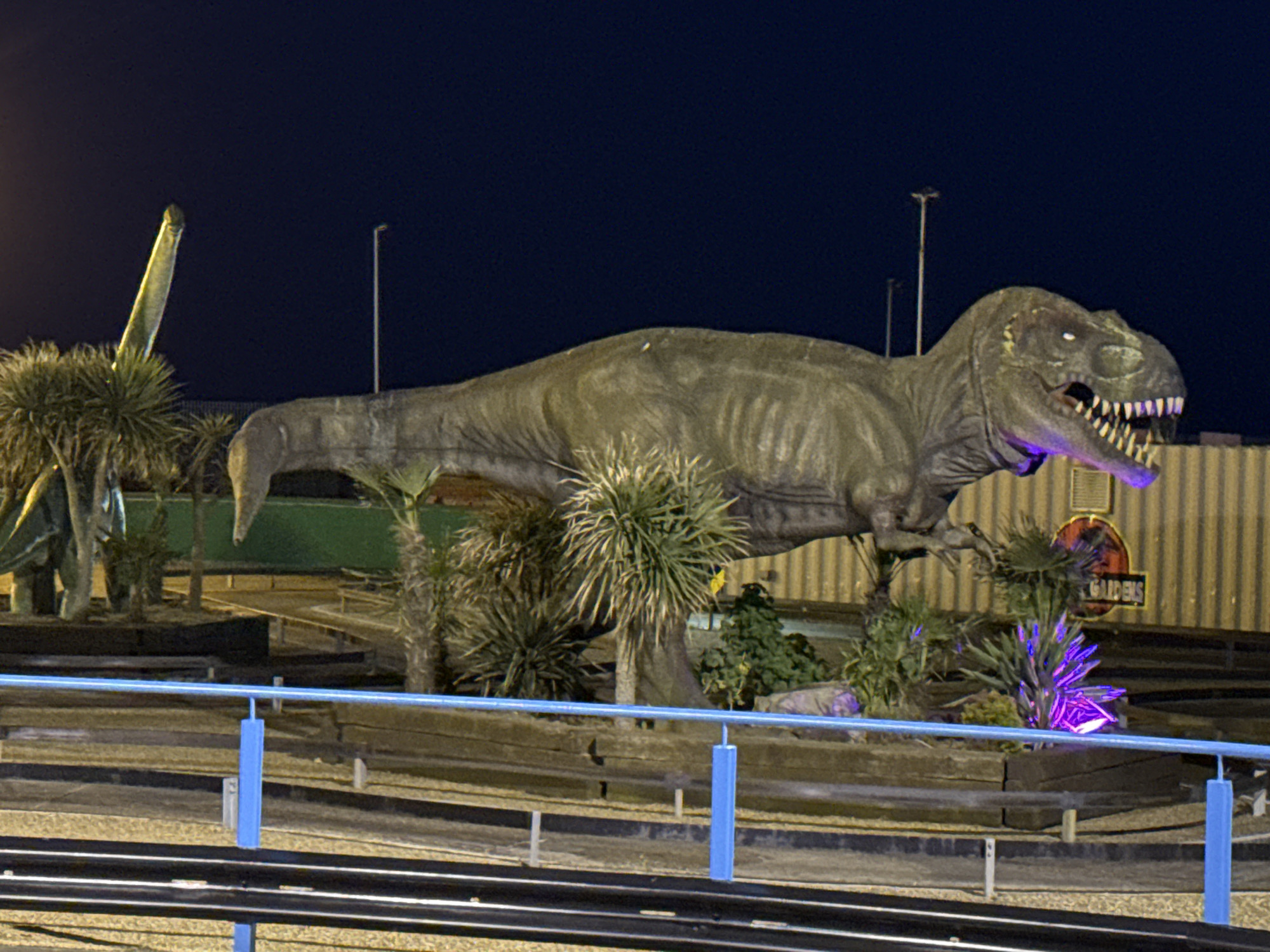 A low-angle, nighttime shot depicts a dinosaur-themed mini-golf course in Gorleston-on-Sea, United Kingdom. The focal point is a large, gray Tyrannosaurus Rex statue with glowing white eyes and purple lighting emanating from its mouth. Behind the T-Rex stands a smaller, green Brachiosaurus statue with a long neck, surrounded by spiky, green yucca plants.
A "Jurassic Gardens" sign is visible next to the T-Rex. The course itself is framed by weathered wooden barriers and blue railings. Railroad tracks run in the lower portion of the frame. Light posts are scattered in the background, illuminating the scene. The overall tone is dark, with the dinosaur statues and course lighting providing the primary sources of illumination.