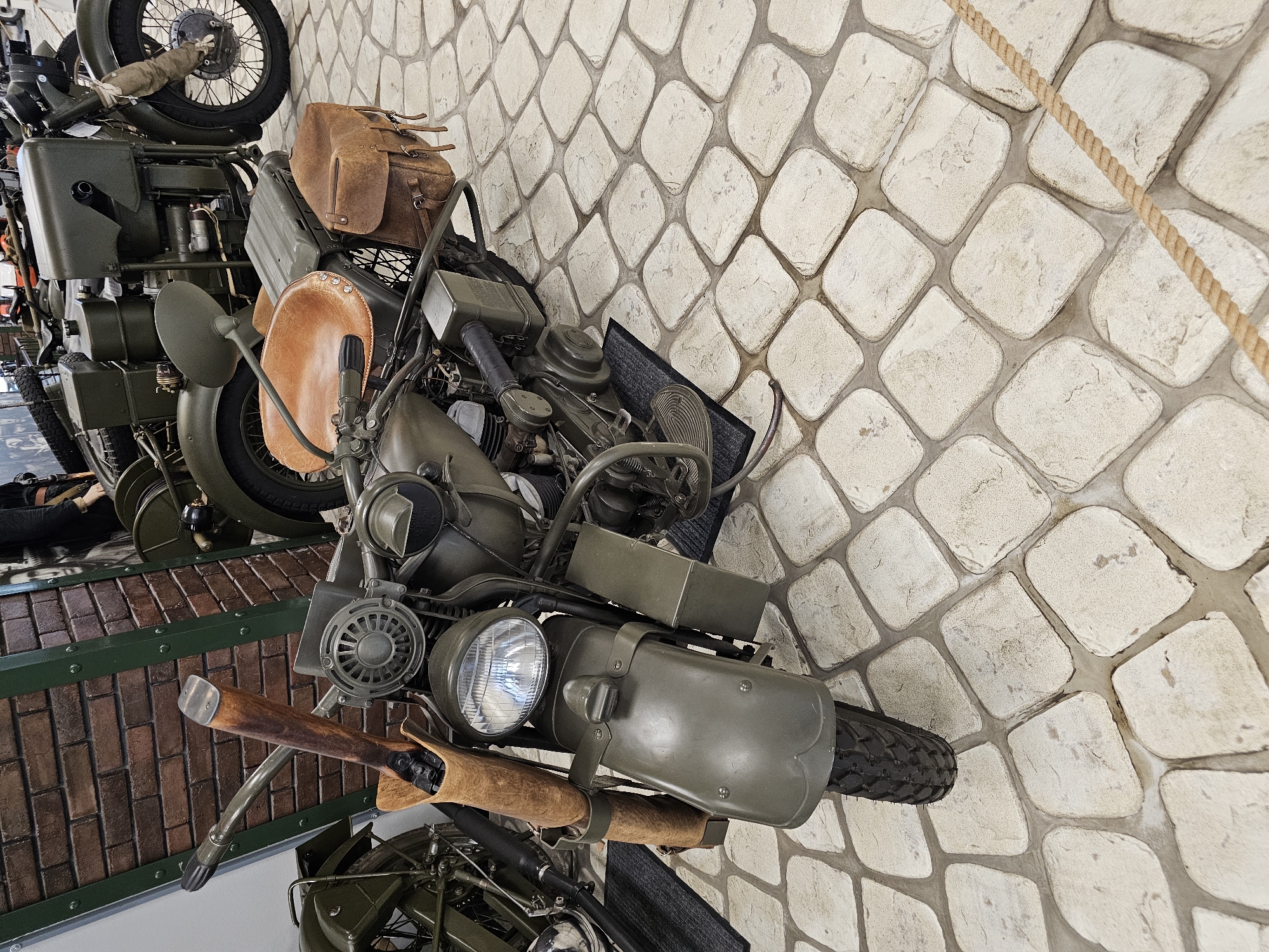 A high-angle shot shows vintage military motorcycles on display in Архангельское, Russia. The motorcycles are painted in drab olive green, with brown leather accents like saddlebags and handlebar covers. The motorcycle in the foreground is the most prominent, revealing detailed engine components, a headlight, and a front fender. Behind, another motorcycle is partially visible, stacked higher than the first. The display area features a faux brick floor with a green-painted border. The backdrop is a wall made of large, irregularly shaped stones in a light beige color. A thick rope, possibly part of a barrier, is visible in the upper right.