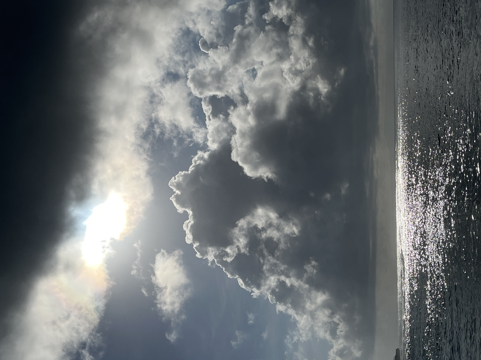 The image shows a view of the sea under a cloudy sky. The water's surface reflects sunlight, creating a shimmering effect. Cumulus clouds dominate the sky, with some areas partially obscuring the sun. The location is likely Kuta Selatan, Indonesia, judging by the coastal setting and the general weather patterns typical of tropical regions. There are no people visible in the shot. The focus is entirely on the natural elements of the sea and sky.