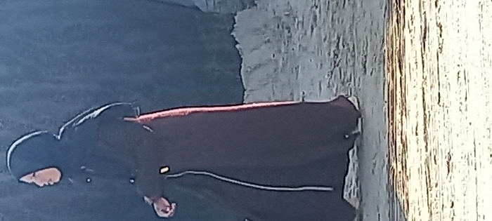 A person in a long, dark garment with a hood stands in front of a textured, light-colored wall. The garment has a burgundy stripe across the chest and a thin white line towards the bottom. The background behind the person is a gradient of dark to light, possibly indicating a transition from shadow to light. The wall features vertical lines and a slightly aged appearance.