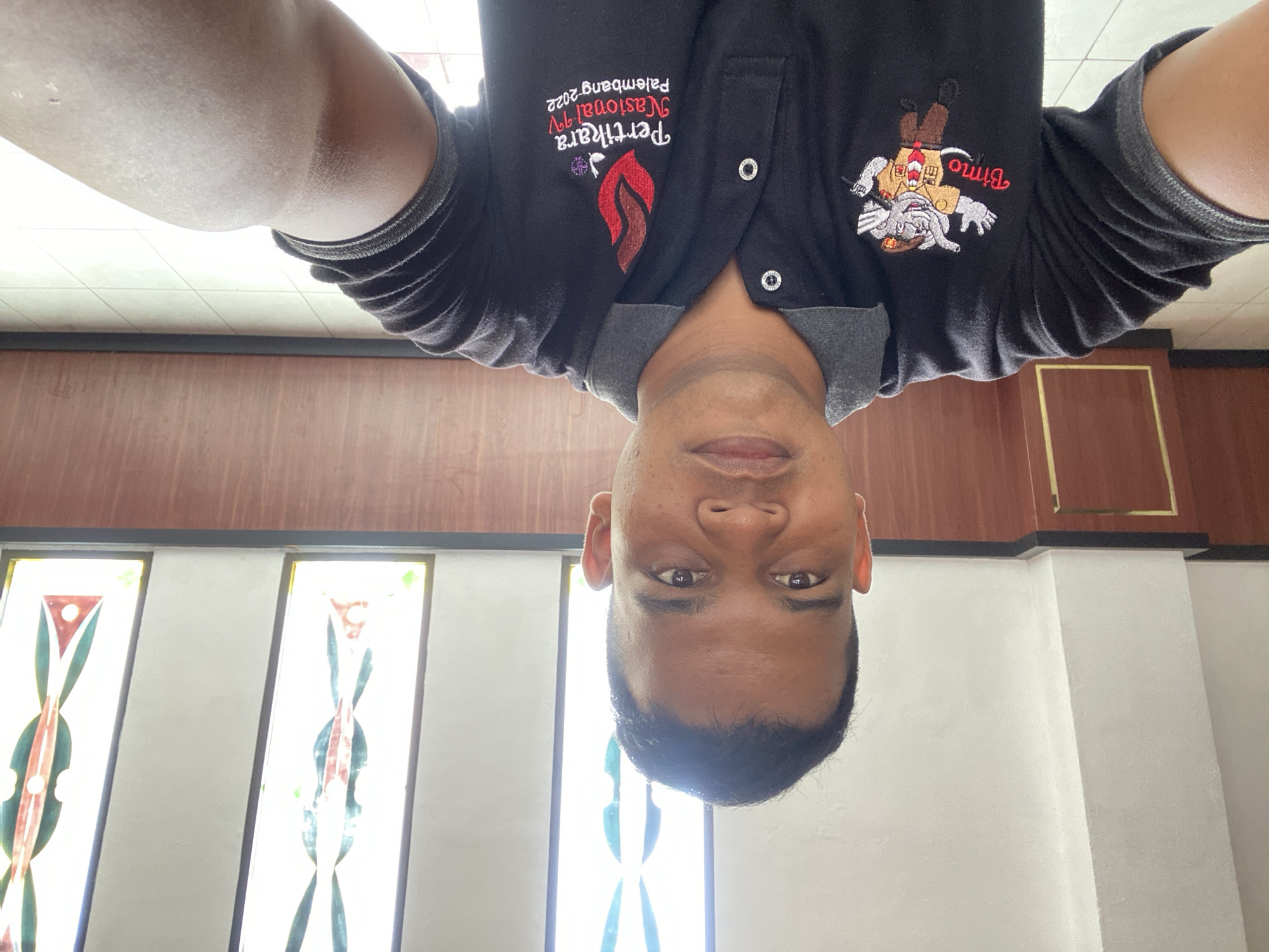 A man is taking a selfie in what appears to be a public building with stained glass windows. He is wearing a black polo shirt with two embroidered logos. The logo on the left features a cartoon elephant in scout uniform with the word "Bimo" written in red cursive. The logo on the right has a flame design and text that reads "Pertikara Nasional IV Palembang 2022". The background shows a wall with wooden paneling and several tall, narrow stained glass windows with geometric patterns in green, white, and red. The floor is tiled. The man has dark skin and short black hair. He is looking directly at the camera. The overall lighting suggests it is daytime. There is no indication of the specific location within the city of Banda Aceh, Indonesia.