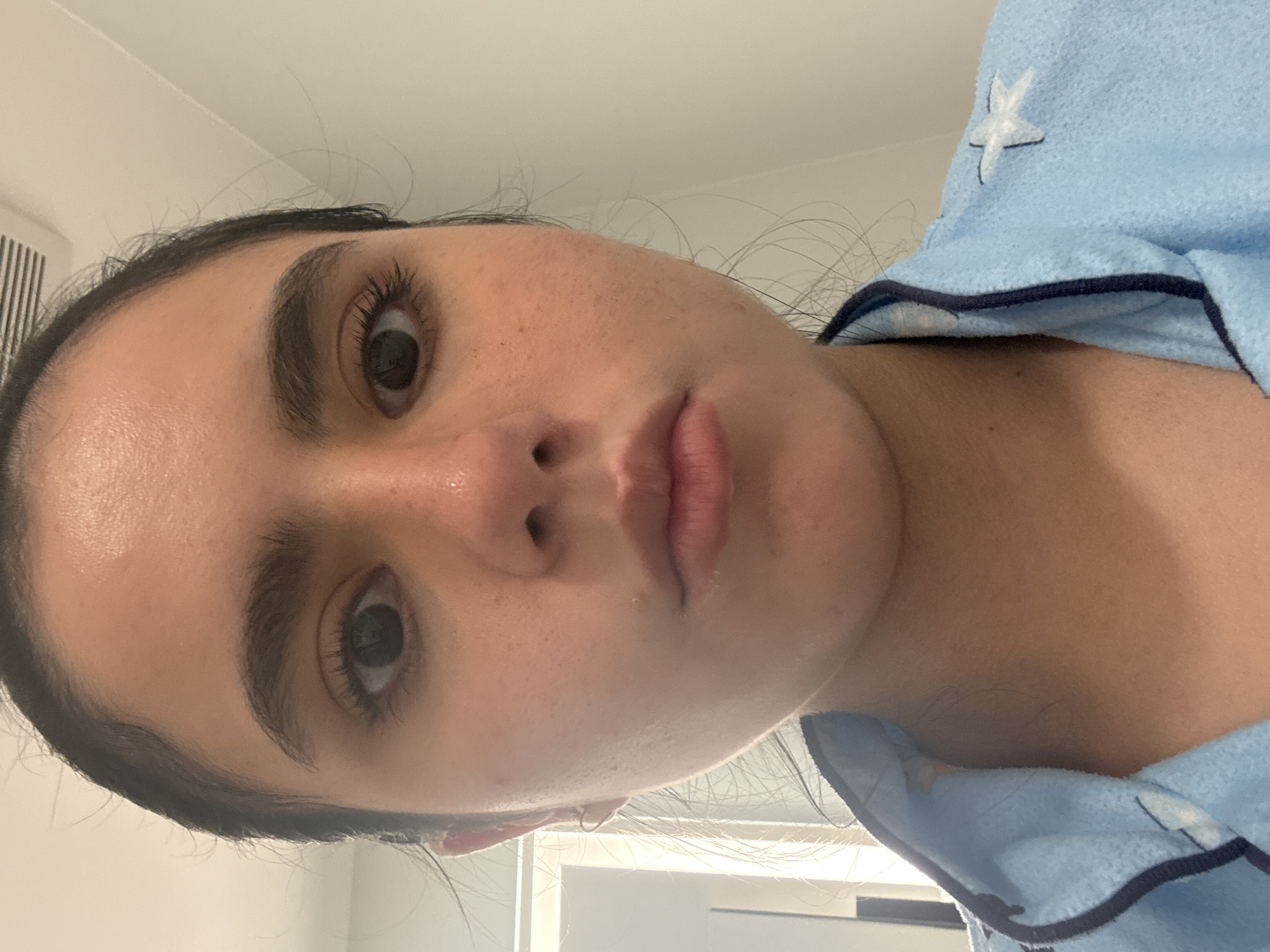 A close-up, head-and-shoulders selfie of a young woman is shown from a low angle, looking directly at the camera. Her dark hair is pulled back. She has dark, thick eyebrows and dark eyelashes. Her eyes are open and dark. Her nose is visible, and her lips are slightly pursed. The woman is wearing a light blue pajama top with a dark blue trim and white star patterns. The background is a plain, off-white wall, with a portion of a white air vent visible at the top center of the image. The lighting is bright, suggesting an indoor environment. The image was taken in Austin, United States of America.