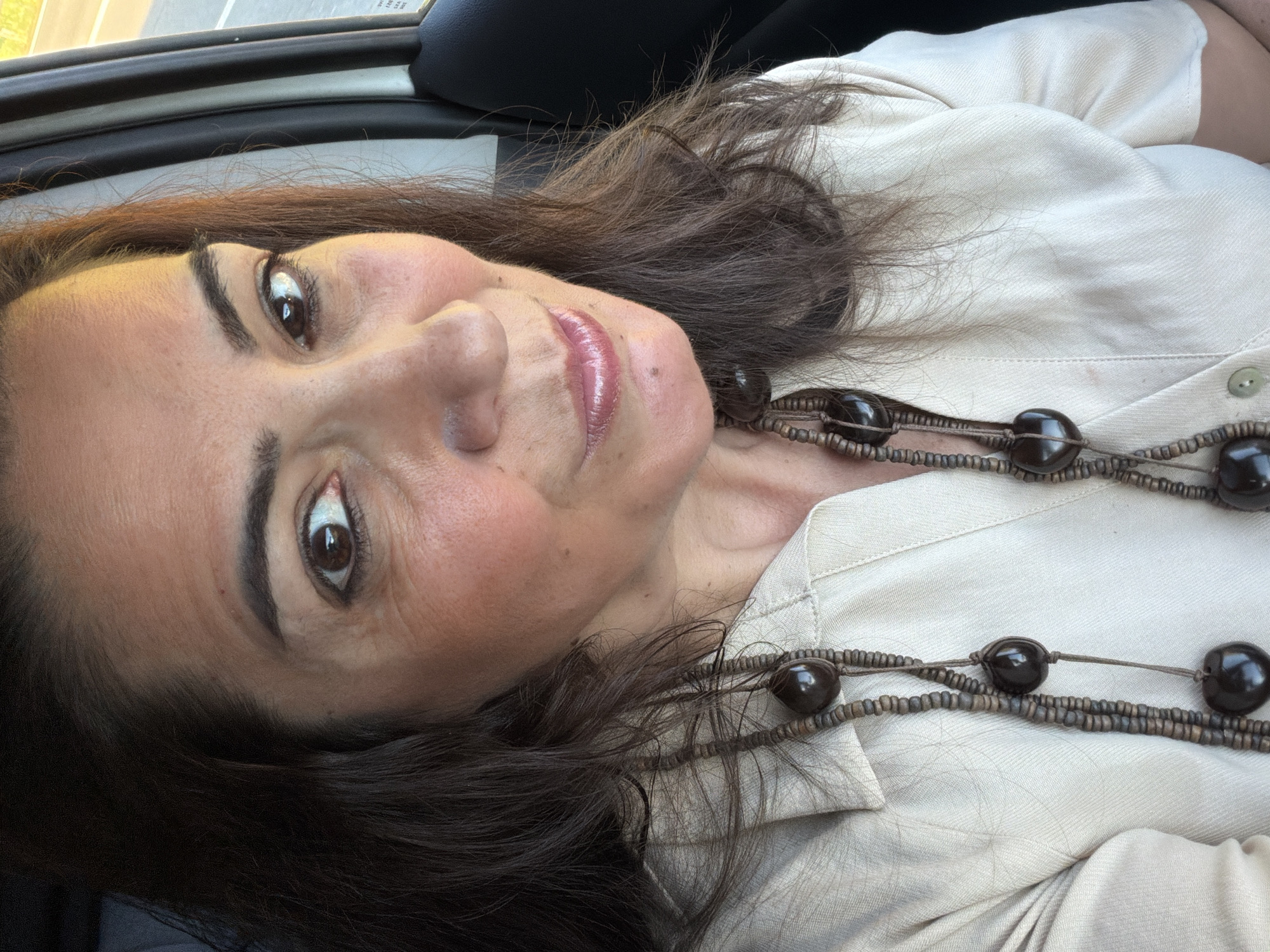 A woman with dark brown hair is facing the camera while inside a car. She is wearing a light-colored, collared shirt and a two-strand necklace made of dark beads. The top of the car window is visible in the upper left corner. No specific details indicate the location is Niederanven, Luxembourg.