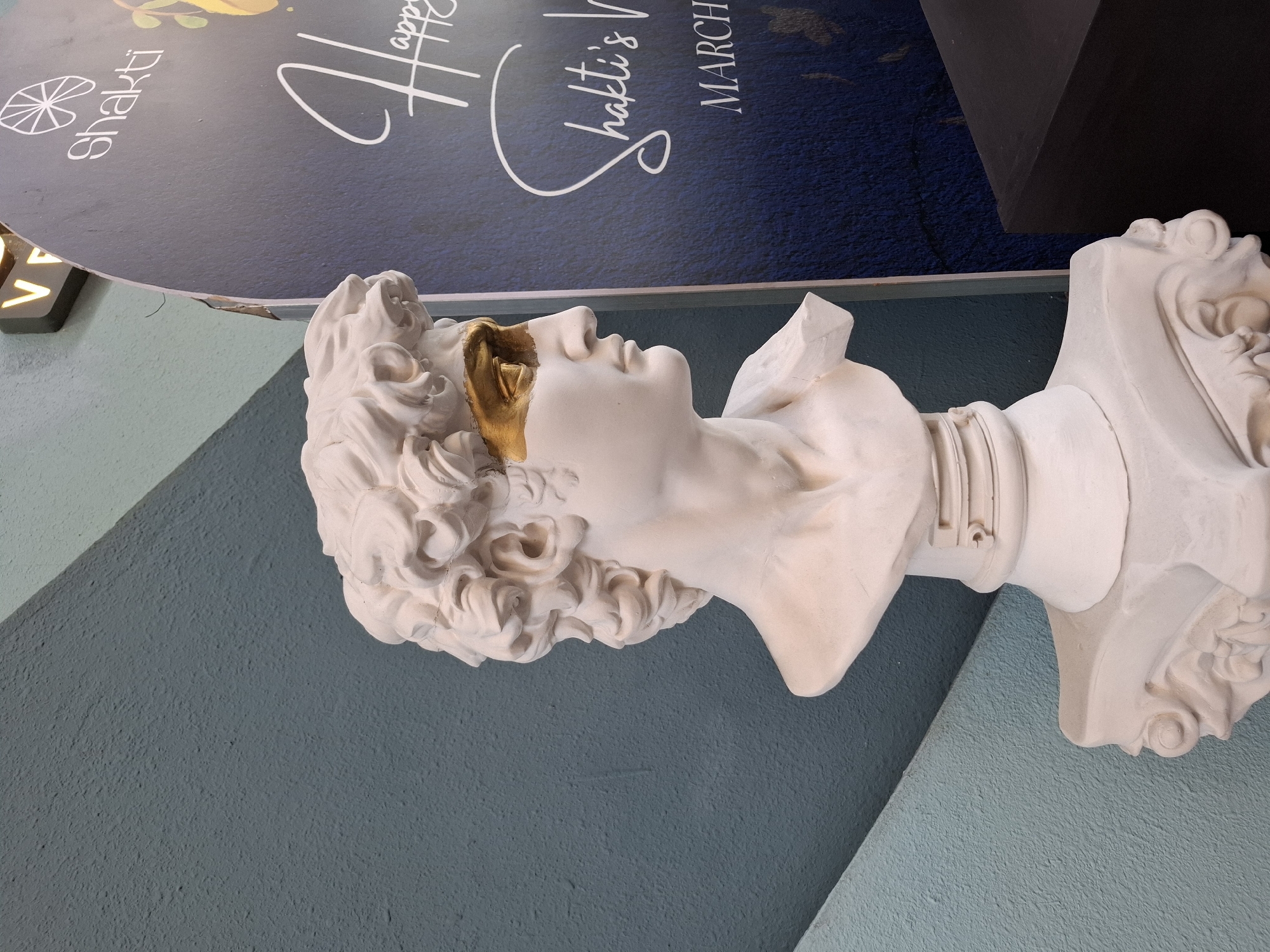 The image depicts a white plaster bust of a man, possibly a replica of a classical sculpture, with a gold-colored accent over its eyes. The bust is positioned on a decorative white pedestal. Behind the bust, a textured teal-colored wall is visible, with a section diagonally receding into a lighter green area. To the right of the bust is a dark blue sign with white text and graphics. The text on the sign includes "shakti," "Happy," and "Shakti's V." Below this, the word "MARCH" is prominently displayed. A dark rectangular object, possibly a piece of furniture, is partially visible in the lower right corner. The lighting suggests an indoor environment, possibly a shop or exhibition space in Nha Trang Ward, Vietnam.