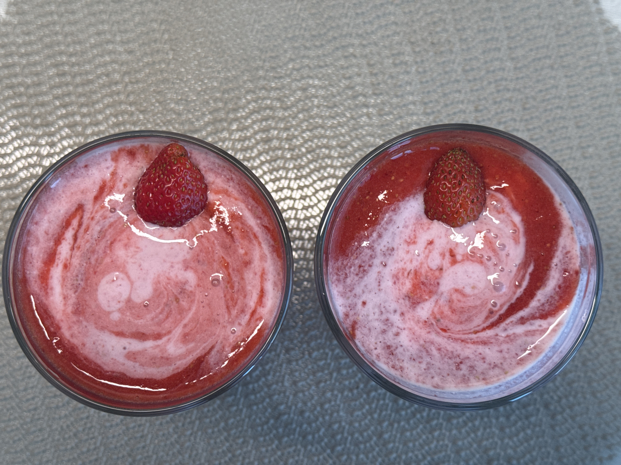 Two glasses of strawberry smoothie are displayed from a top-down view. The smoothies have a creamy texture with swirls of pink and white, topped with whole strawberries. The glasses are clear and sit on a textured, light-colored surface, possibly a table. The background is out of focus, but appears to be a woven placemat or similar material. The lighting suggests it is daytime. No people or text are visible. The overall impression is a refreshing beverage, likely taken outdoors or in a well-lit room.