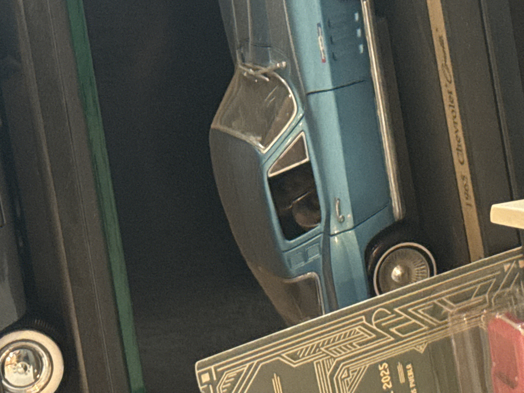 The image shows a close-up of what appears to be a die-cast model car, specifically a 1963 Chevrolet Corvette. The model is light blue and is presented in its packaging. The location is implied to be in the city of Puebla, Mexico, based on a partial view of what seems to be packaging related to the year "2025" and "Puebla". The background is dark and out of focus.