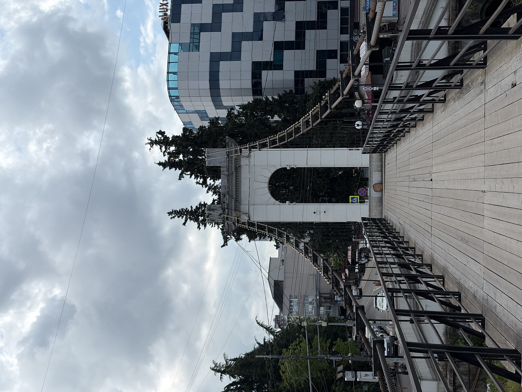 The image depicts a pedestrian bridge in Sochi, Russia. The bridge has a wooden deck and is flanked by black metal railings. In the center of the bridge, a large white arch structure supports the suspension cables of the bridge. Behind the arch, several tall, dark green evergreen trees are visible. To the right of the trees, a modern building with a white facade and blue glass windows stands prominently. The sky is overcast with gray clouds. In the background, other buildings and cars are visible along a street. The scene is captured from the perspective of someone standing on the bridge, looking forward.