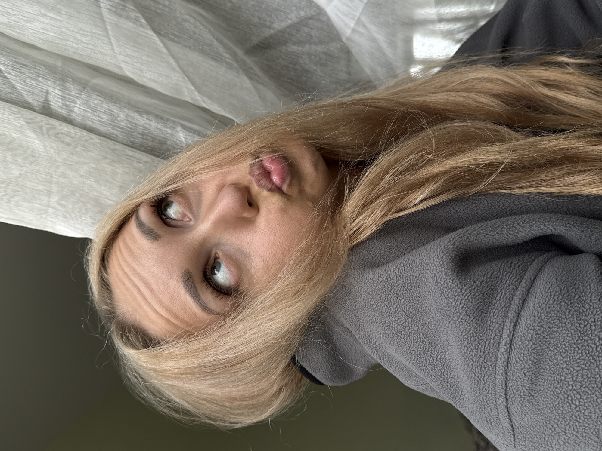 This image, captured in an indoor setting in the city of Chaikovsky, Russia, features a light-skinned individual, likely female, with long, straight blonde hair. She is wearing a grey fleece garment with a visible black stand-up collar. Her light-colored eyes are directed upwards and slightly to the viewer's right, an expression that causes several horizontal lines to form on her forehead. Her lips are tightly pursed into a "duck face" or "fish face" pose. The immediate background to the upper left is a solid dark surface, while the upper right features a light-colored, vertically textured fabric, consistent with a curtain or drape.