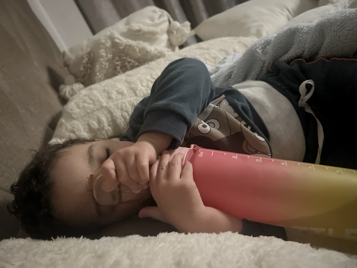 A toddler lies asleep on a plush, light-colored sofa, nestled amidst soft pillows and a blue blanket. The child wears a patterned long-sleeved shirt and dark shorts. One hand clutches a clear pacifier near their face, while the other rests against a large, gradient-colored water bottle. The overall scene suggests a moment of peaceful rest and comfort.