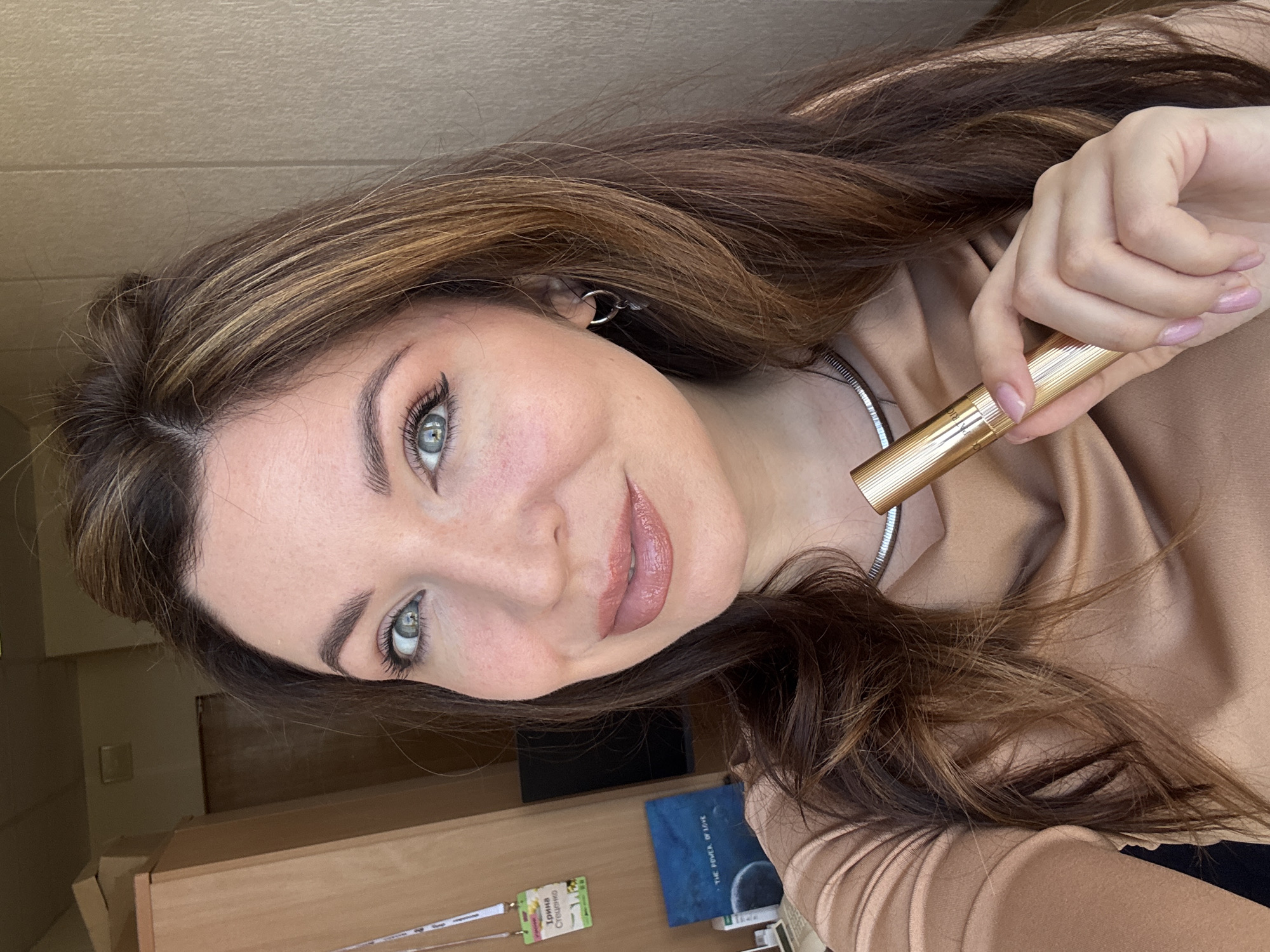 A woman is holding a gold tube in her right hand. She has brown hair with blonde highlights, fair skin, and light-colored eyes. She is wearing eye makeup including eyeliner and a lip color that matches her skin tone. The background includes a wooden surface, a blue object with the text "THE POWER OF LOVE" visible, and a lanyard with a badge. The badge contains text that includes "Юлия Стрещенко", which translates to "Yulia Streshchenko", indicating a possible name. There is no confirmed information about the location in Kryvyi Rih, Ukraine.