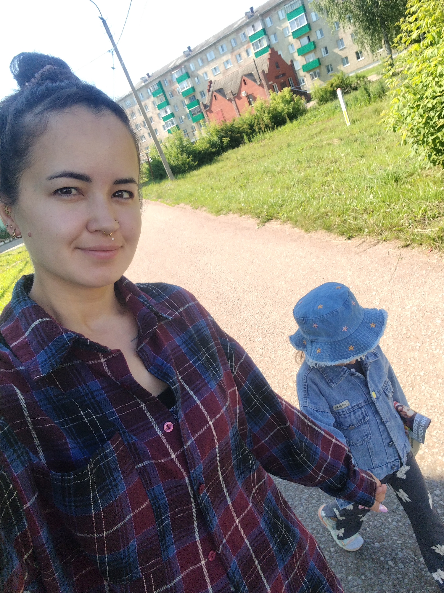 A woman with dark hair pulled into a bun and facial piercings is taking a selfie outdoors. She is wearing a plaid shirt. Beside her, a young child wearing a denim bucket hat and denim jacket is walking away from the camera, holding the woman's hand. They are on a paved path in a grassy area with trees and multi-story buildings in the background. The buildings appear to be residential. This scene is likely located in Yanaul, Russia, given the context of the request.