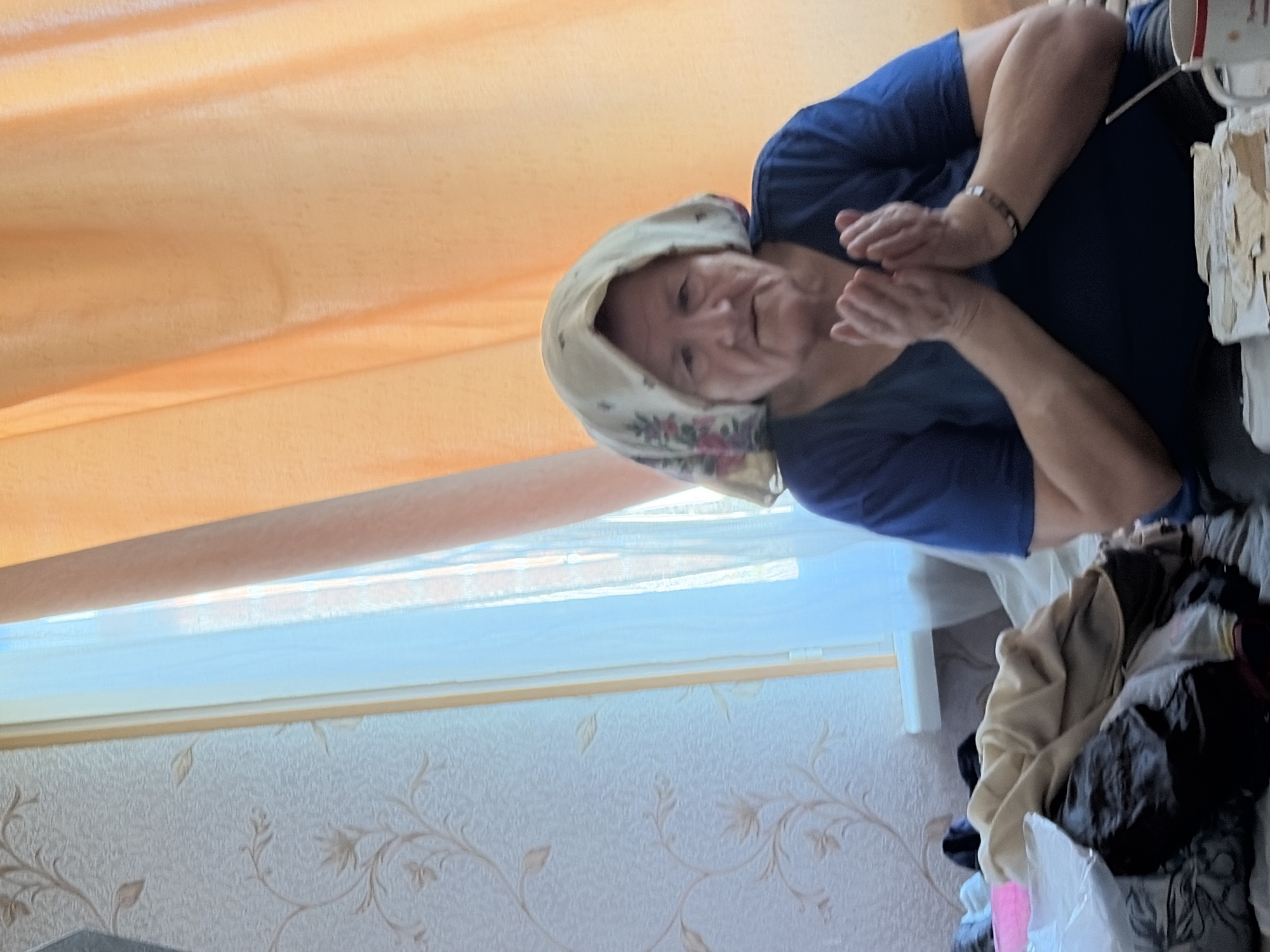 An elderly woman with a floral-patterned headscarf over her hair and a dark blue v-neck shirt is sitting. Her hands are clasped together in front of her chest. She has a neutral expression. The background shows a wall with patterned wallpaper on the left and orange curtains on the right. There is a window with sheer blue curtains in between. A pile of folded clothes is visible in the foreground to the left of the woman. The scene appears to be indoors, possibly in a residential setting in Tambov, Russia.