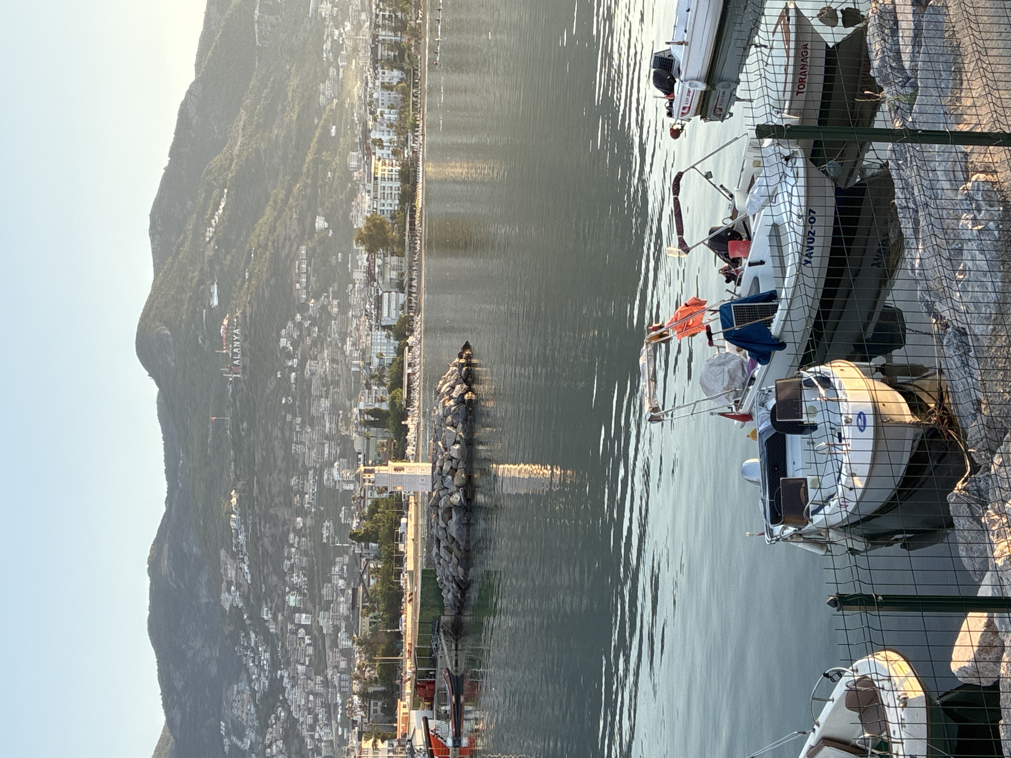 The image depicts a calm harbor in Alanya, Turkey, with a backdrop of a densely populated hillside covered in buildings and lush greenery. In the foreground, several small boats are moored along a rocky shore, separated by a chain-link fence. The boats are white with dark trim, and some have colorful flags and awnings.
A prominent white lighthouse, rectangular in shape with decorative patterns, stands on a rocky island in the middle of the harbor. Its reflection is visible in the still water. In the distance, a long stretch of beach is lined with buildings and palm trees, suggesting a resort town. The mountain range behind the city is a significant feature, with structures and a prominent "ALANYA" sign visible on its slopes. The sky is clear and bright, indicating daytime, likely morning or late afternoon due to the soft, warm light. The overall scene conveys a sense of tranquility and scenic beauty.