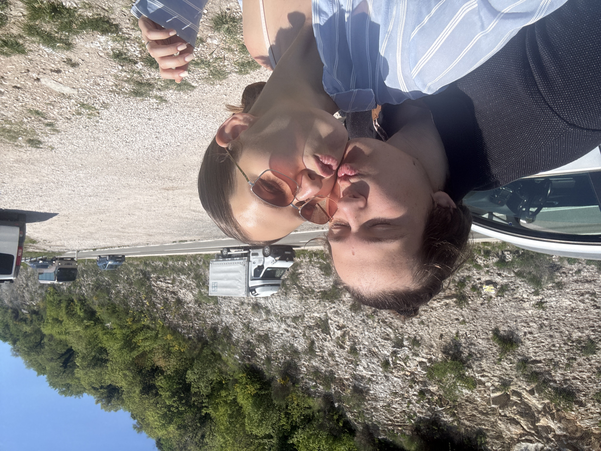 This image, correctly oriented, depicts two individuals, a woman and a man, engaged in a kiss by a roadside in Stanišići, Montenegro. The woman, on the left, wears sunglasses with pink-tinted rectangular frames and a blue and white striped long-sleeved shirt over a lighter top. A ring is visible on her left ring finger. The man, on the right, also wears a blue and white striped shirt beneath a dark, possibly navy or black, jacket with a subtle dotted texture. Their faces are close, lips pressed together in an affectionate gesture.
The background reveals an outdoor setting on a bright, sunny day. Immediately behind the couple is a gravelly or dirt area with sparse green growth, leading to a paved road. Several vehicles are visible on the road, including a prominent white cargo van, a dark SUV, and other cars, suggesting a well-traveled route. Beyond the road, a steep, rugged hillside rises, densely covered with green trees, bushes, and exposed rock formations, characteristic of a mountainous or hilly landscape. A clear blue sky is visible towards the lower left. The lighting indicates strong daylight, with clear shadows. No discernible text is visible on any objects or surfaces within the image. The overall impression is one of intimacy and affection amidst a scenic, natural environment.