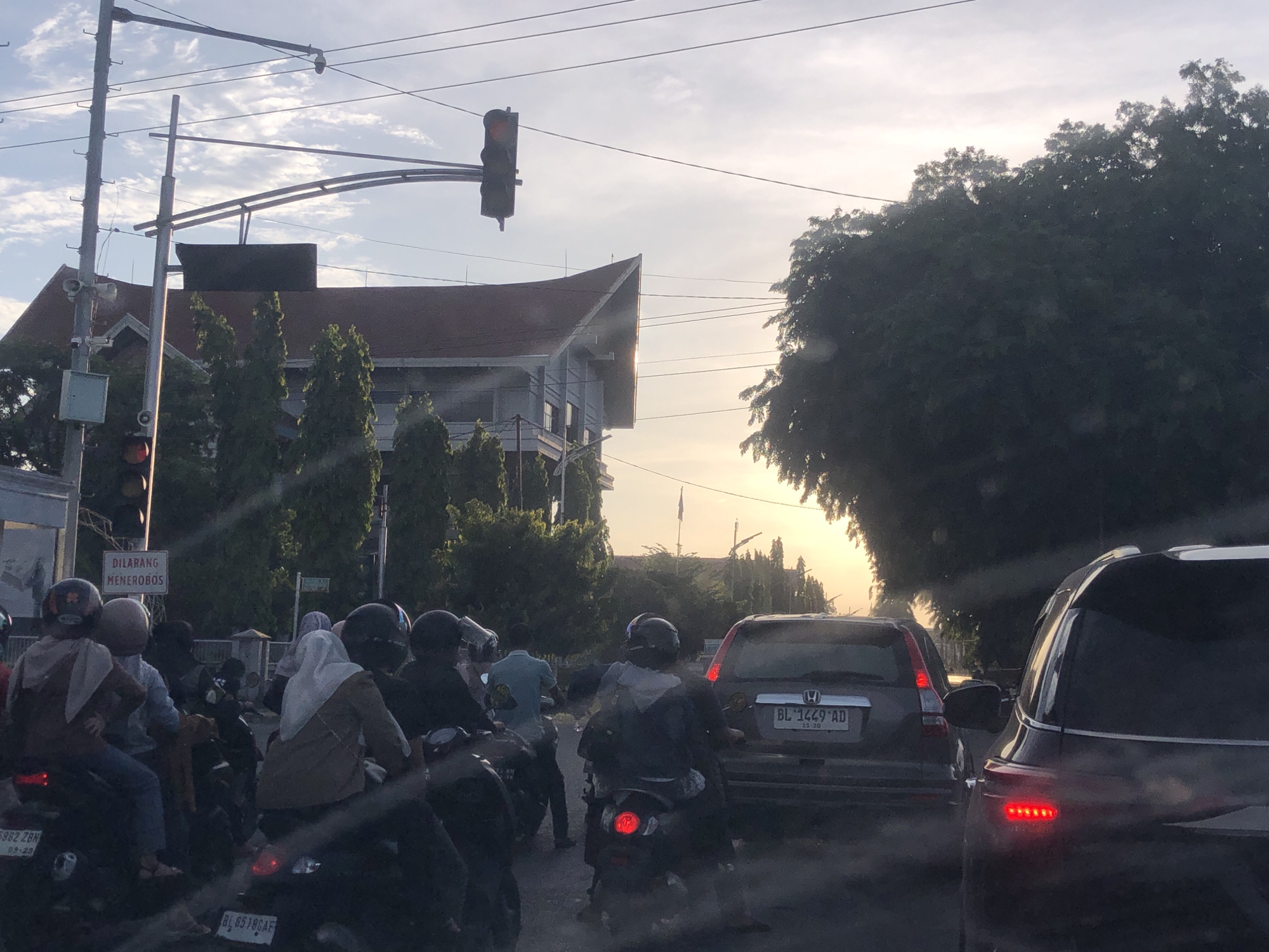 The image shows a traffic scene in Lhokseumawe, Indonesia. A group of people on motorbikes and cars are stopped at a traffic light. The light is red. A building with a multi-level facade is visible behind the traffic lights. The sky is bright with the sun setting or rising in the distance. A sign that says "DILARANG MENEROBOS" is seen. The license plate on a grey car says "BL 1449 AD".