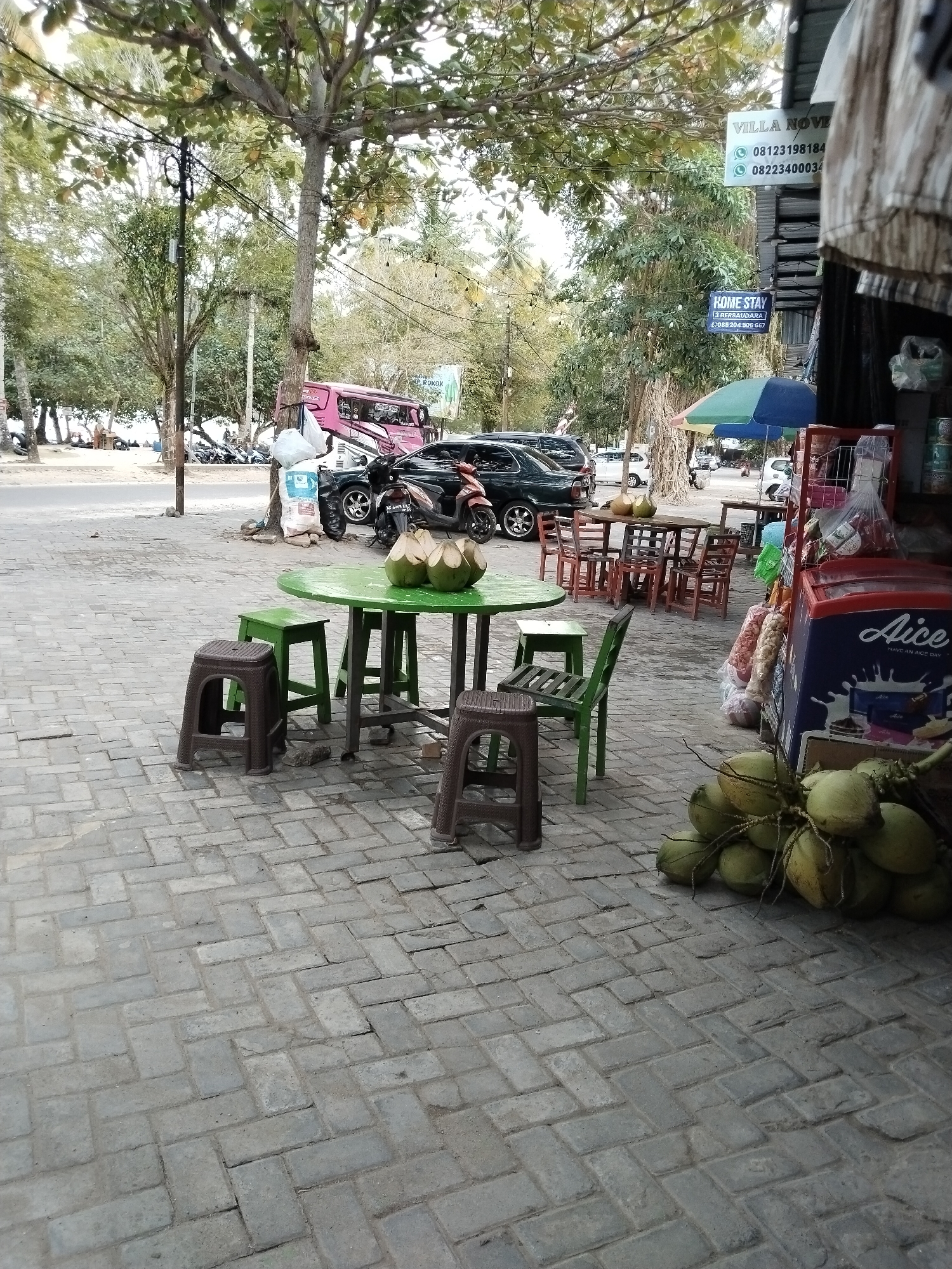 The image captures a scene in Prigi, Indonesia, likely a roadside area near a beach or tourist spot. In the foreground, a green circular table is set with four brown plastic stools and two green stools, one of which is partially obscured. Three coconuts are placed on the table. A cluster of green coconuts sits on the paved ground to the right.
Behind the table, a small eatery or shop is visible, featuring a red and blue refrigerator with "Aice" branding. Bags of snacks and other items are displayed outside the shop.
In the background, a pink van is parked, and several cars and motorcycles are visible on the road. A street umbrella, partially open, stands near the shop. Trees with lush foliage line the street, and in the distance, there appears to be a sandy area, possibly a beach, with people lounging. Overhead, power lines and streetlights are present. Signs for "VILLA NOVE" and "HOME STAY" are mounted on a pole.