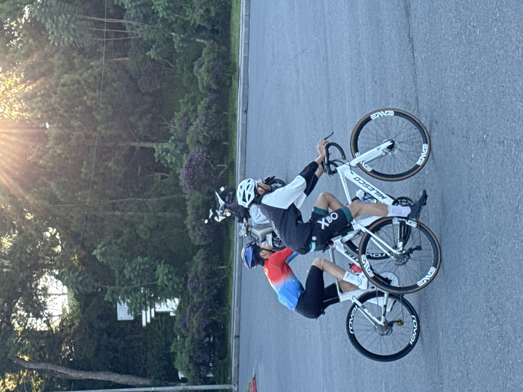 Two individuals are cycling on a paved road in Da Lat, Vietnam, with a lush green forest and purple flowering bushes in the background. The cyclist in the foreground is wearing a white helmet, a black and white cycling jersey, and patterned shorts. The cyclist behind them is wearing a blue and red cycling jersey and black shorts. Both cyclists are on white road bikes with prominent brand names visible on the wheels. In the background, two motorcycles are parked, and the sun's rays are filtering through the trees, creating a dappled lighting effect. The overall scene suggests an outdoor recreational activity in a scenic, natural setting.